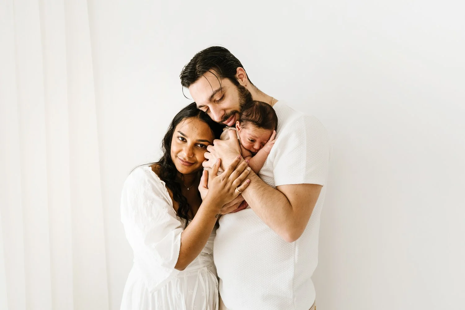 A diverse family of three, including a woman, a man, and a newborn baby, standing against a plain white background, sharing a tender moment of love and bonding.