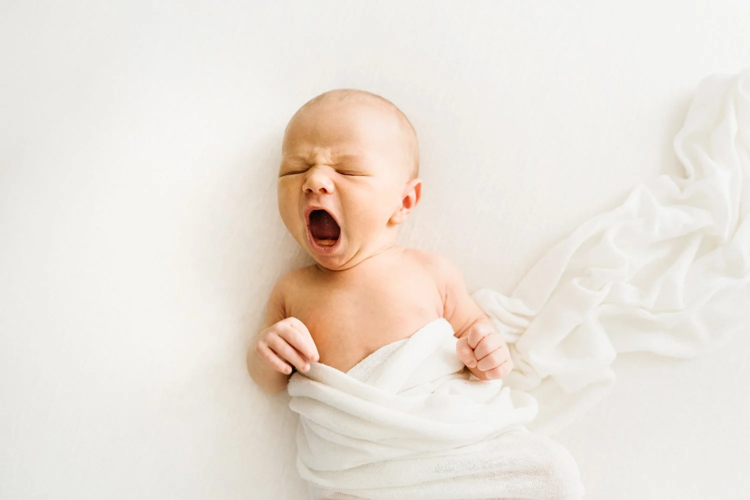 A newborn baby yawning while lying on a white surface, wrapped in a white cloth.