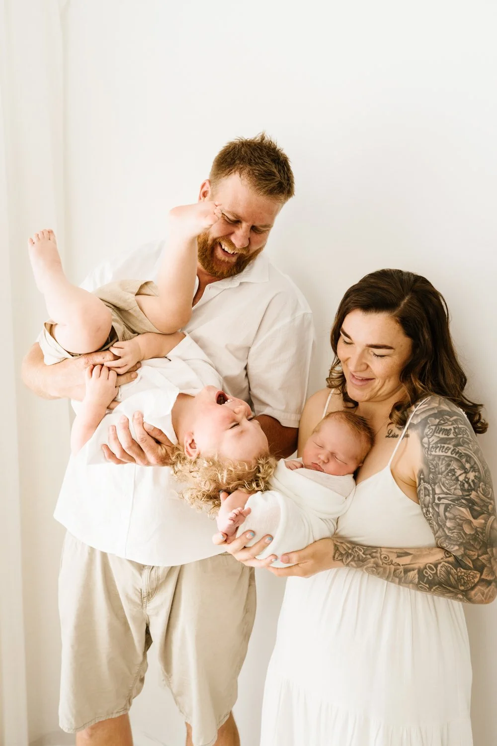 A couple with two young children, one holding a newborn, smiling and enjoying a family moment in a bright room.
