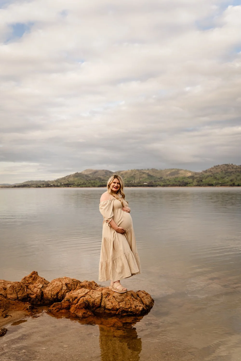 A pregnant woman wearing a beige dress standing on rocks in a calm body of water, smiling with hills and a cloudy sky in the background.