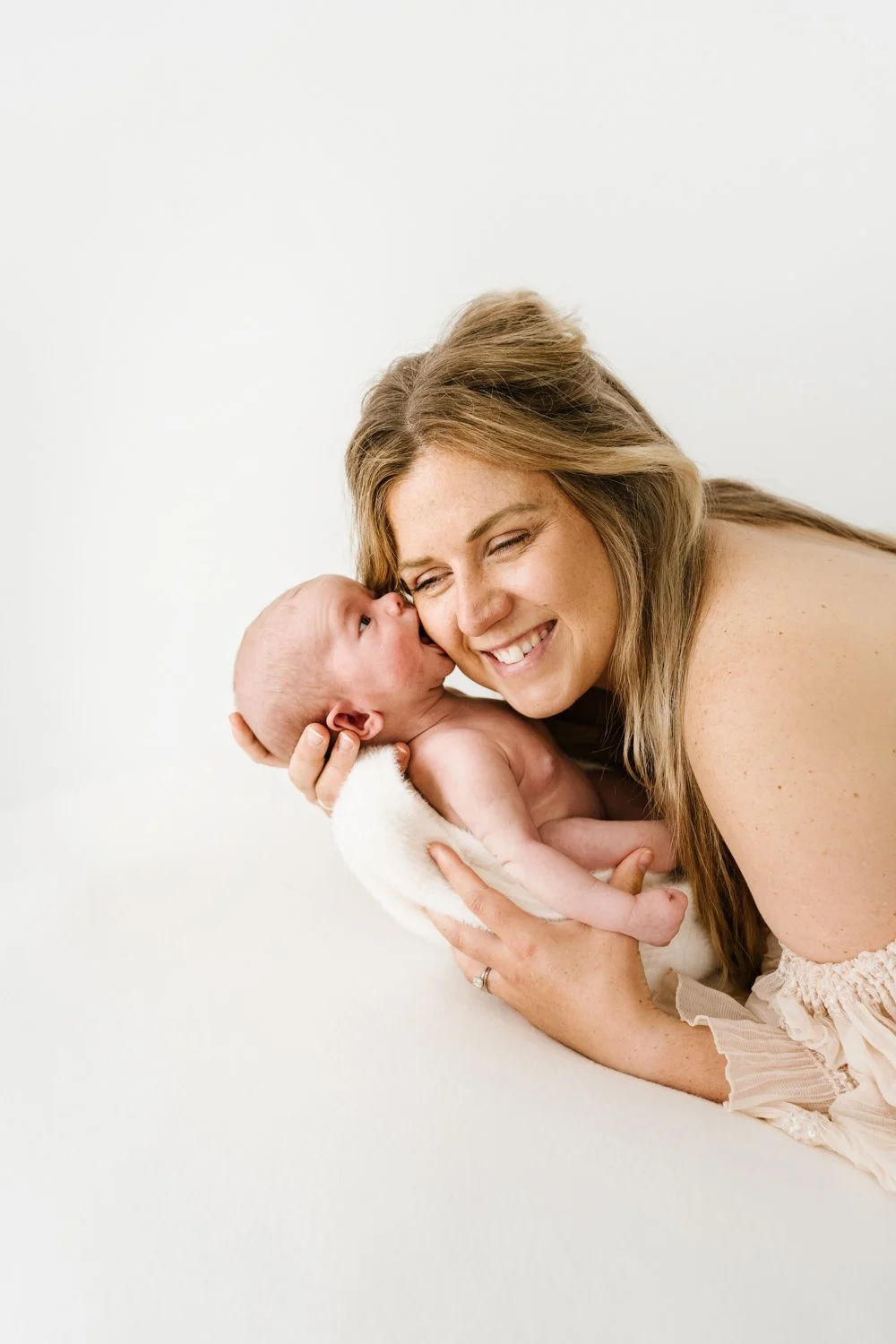 A woman holding a newborn baby close to her face, smiling with her eyes closed, in a bright, neutral background.