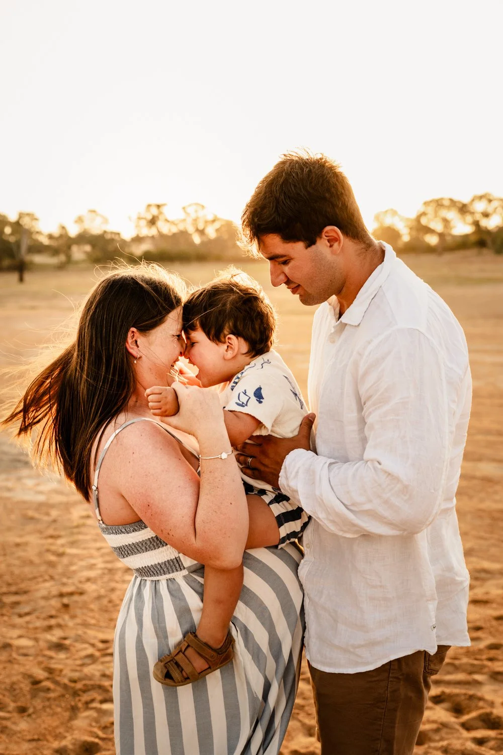 A family of three standing outdoors during sunset, with the mother holding the young boy, and the father looking at them, all sharing a tender moment.