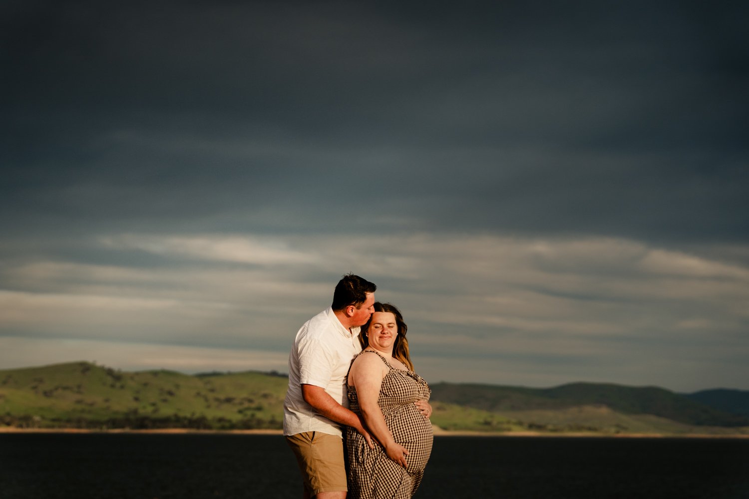 A couple, with the man kissing the woman on the head, standing outdoors near a body of water with green hills under a dark, cloudy sky.
