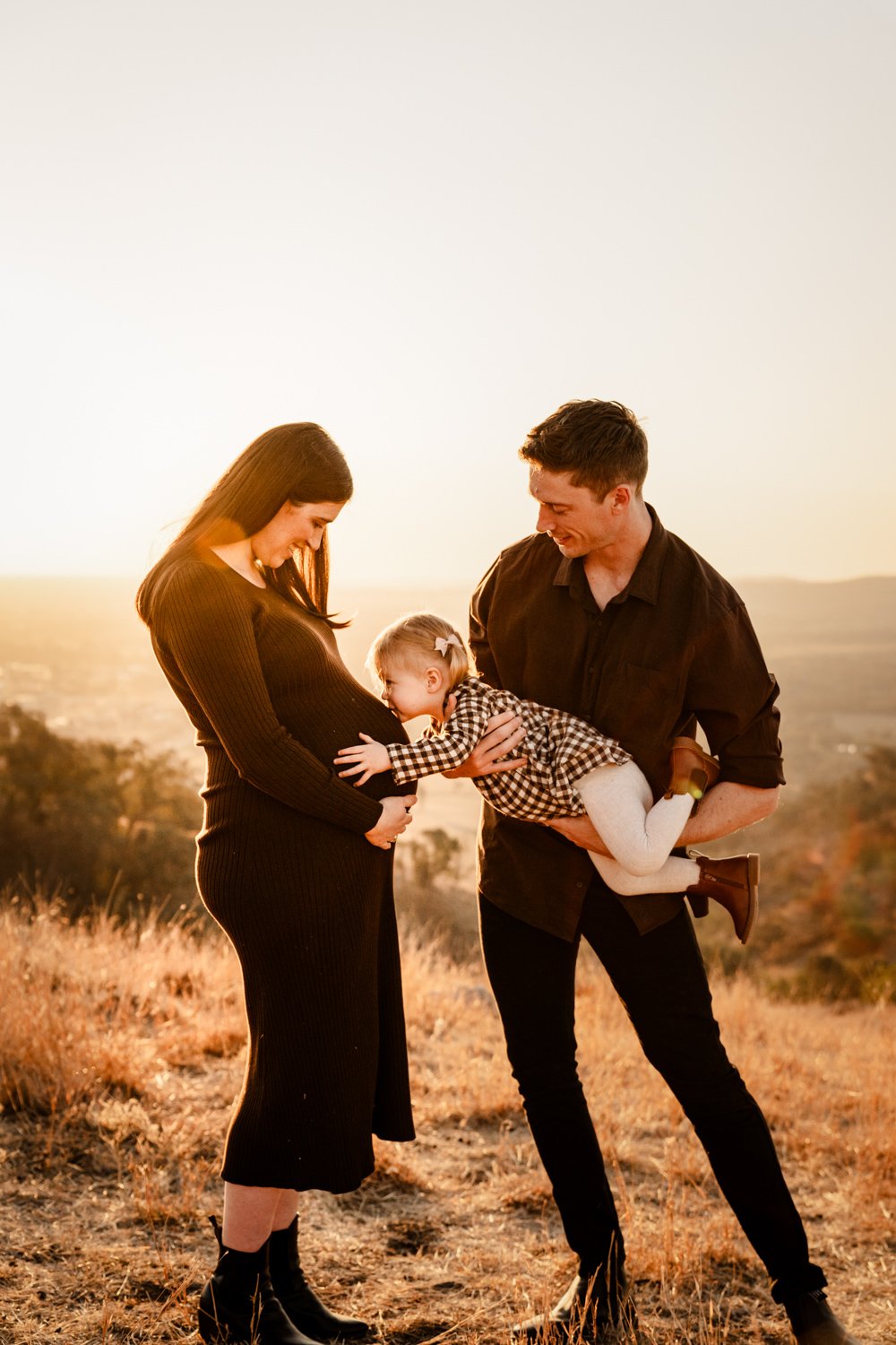 A family of three outdoors during sunset, with a pregnant woman in a black dress, a man holding a small girl in a checkered dress, all smiling.