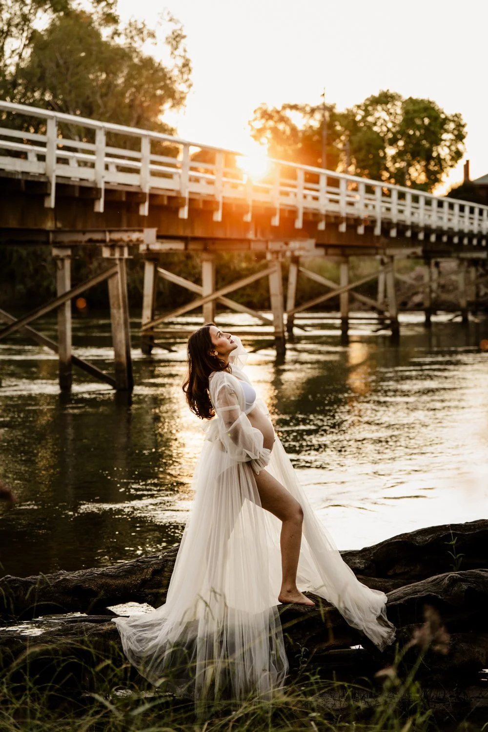 A woman in a long, flowing white dress stands on rocks by a river during sunset, with a bridge and trees in the background.