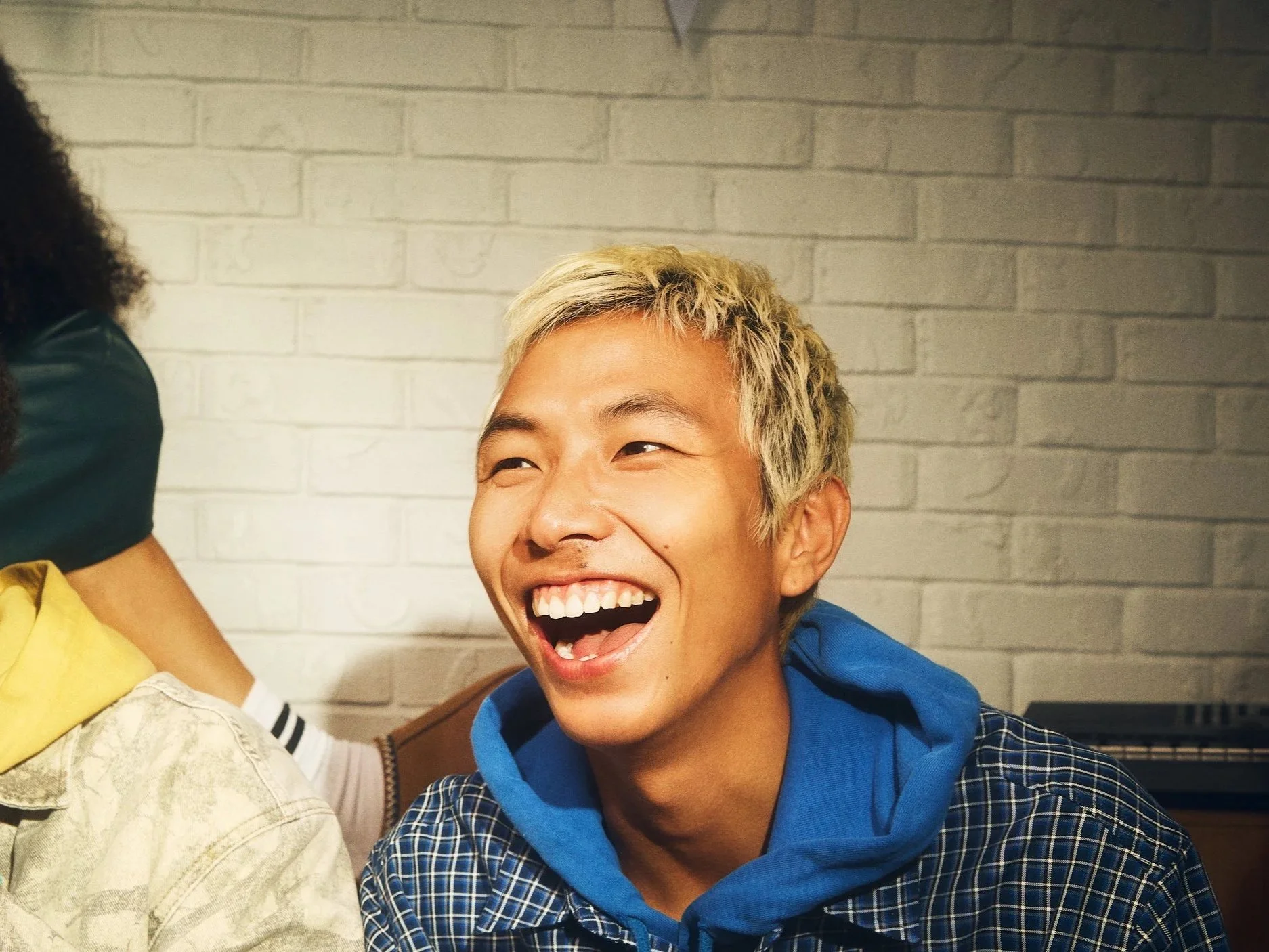 A young man with platinum blonde hair, wearing a blue hoodie and checkered shirt, laughing with his mouth open against a white brick wall.