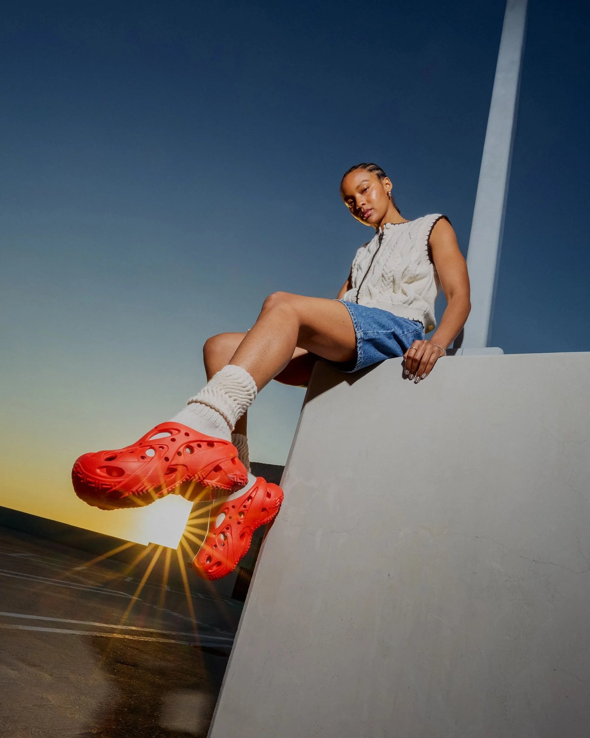 Young woman wearing a sleeveless white vest, blue shorts, and white socks with bright orange slip-on shoes, sitting on a ledge with a sunset and a steel pole in the background.