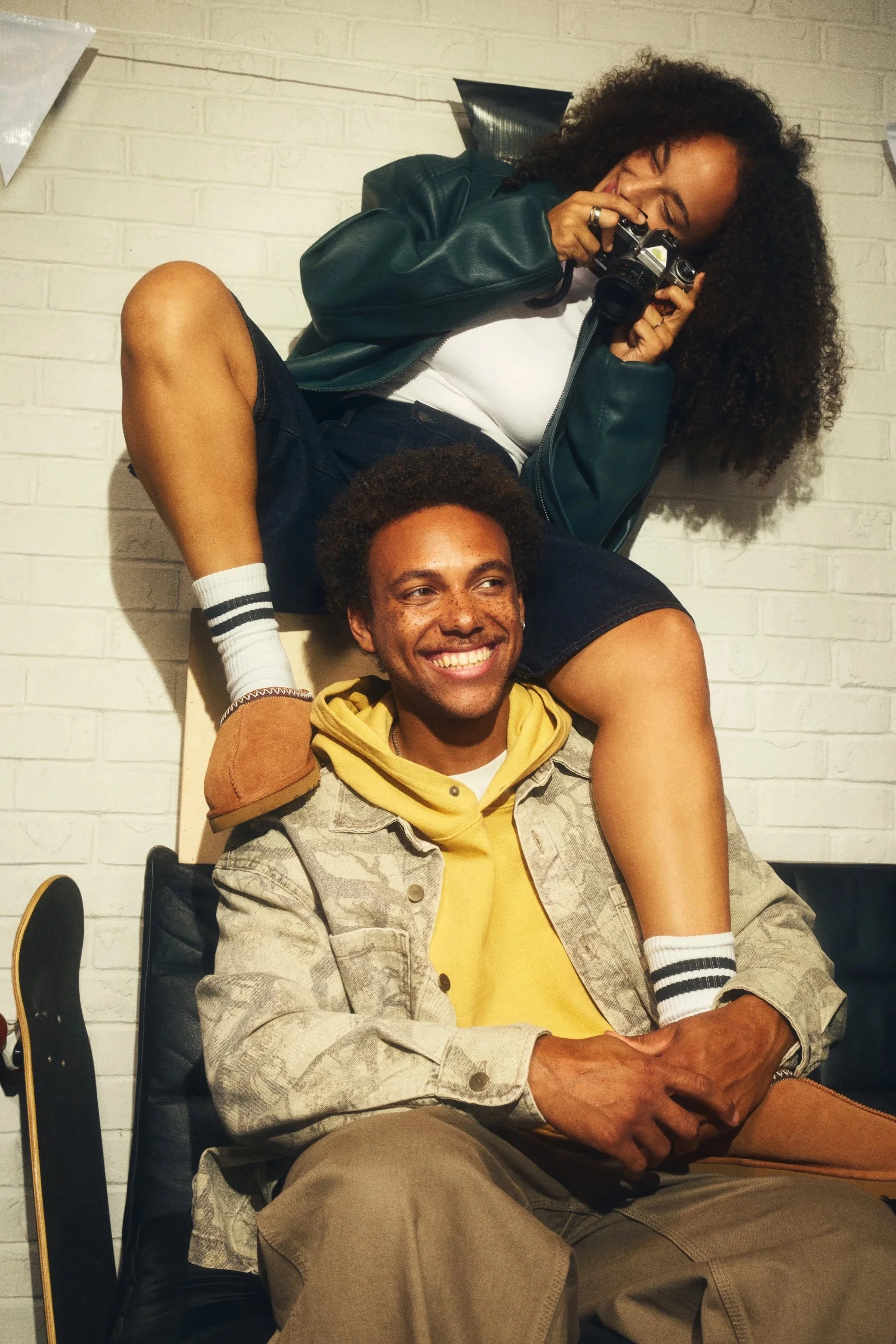 A woman with curly hair is sitting on a person's shoulders, taking a photo with a camera. The person sitting is smiling and wearing a yellow hoodie and beige pants. They are in front of a white brick wall.