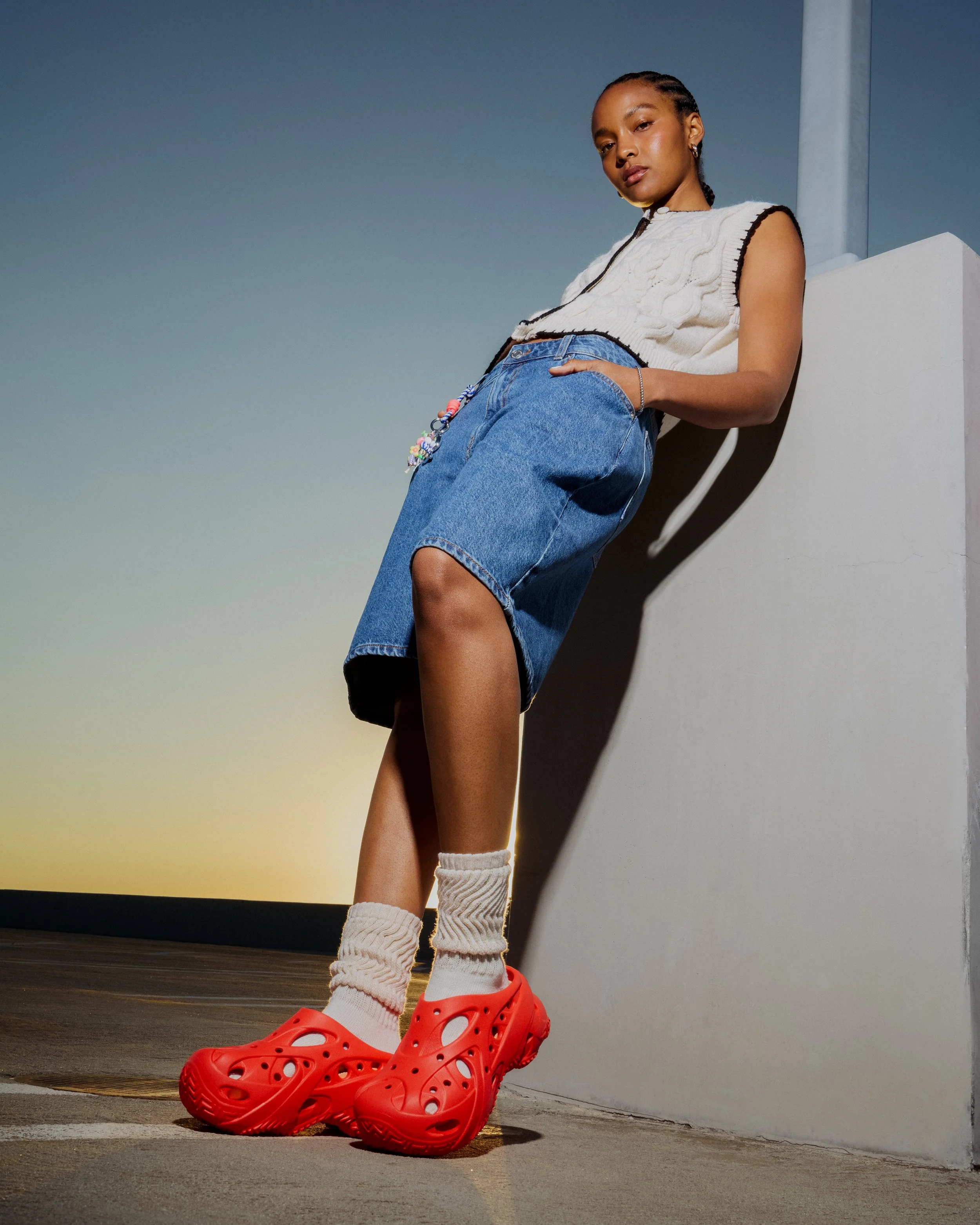 Young woman wearing a sleeveless light-colored sweater, denim skirt, white socks, and red Crocs posing outdoors near a wall during sunset.