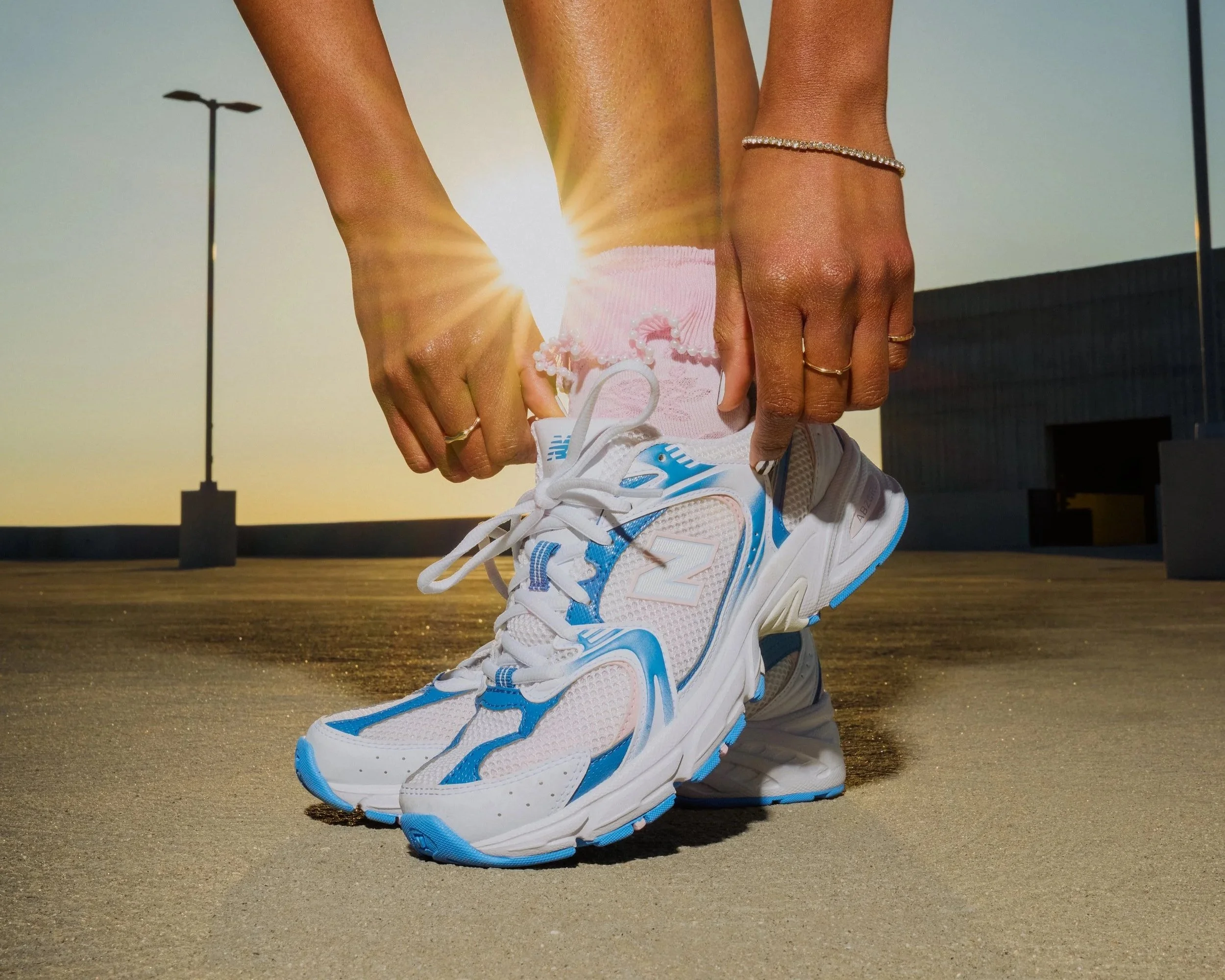 A person wearing a white and blue athletic shoe, pink socks, and jewelry is tying their shoelaces outside on a concrete surface during sunset, with the sun shining in the background.