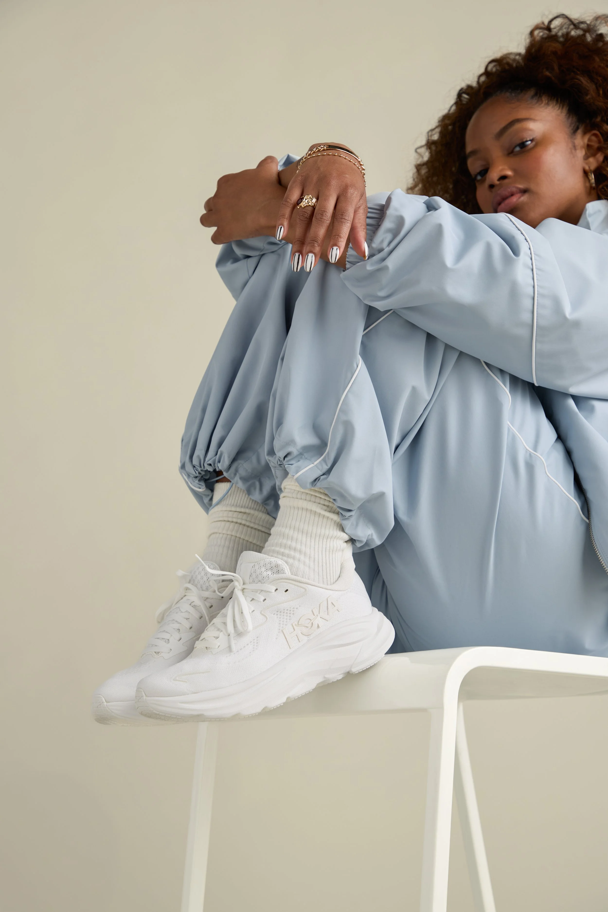 A woman sitting on a white chair, wearing a light blue jacket, white sneakers, and beige socks, with her legs crossed and arms resting on her knees.