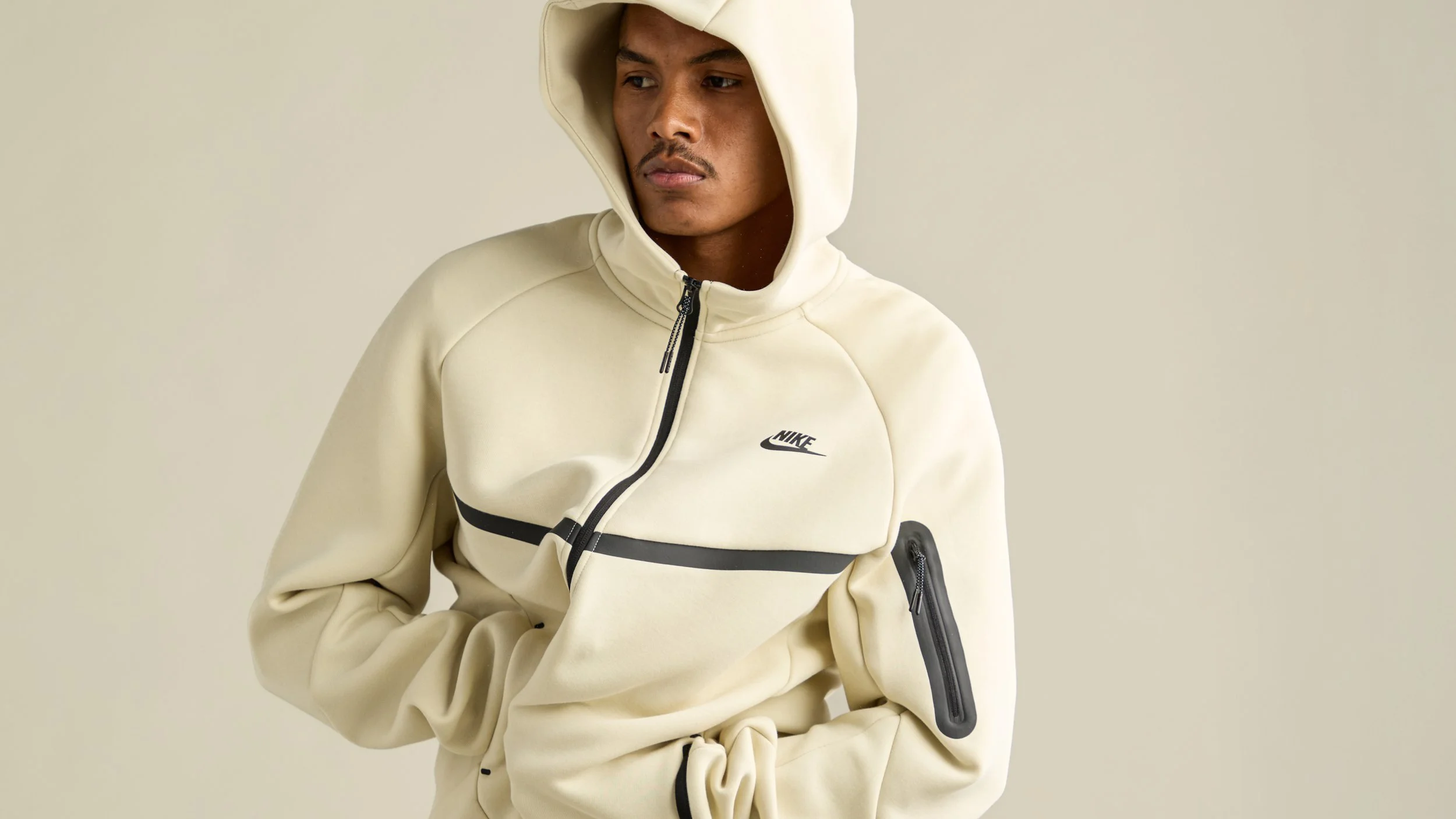 A young man wearing a beige Nike hoodie with a hood, black zipper, and black accents, standing against a plain neutral background.