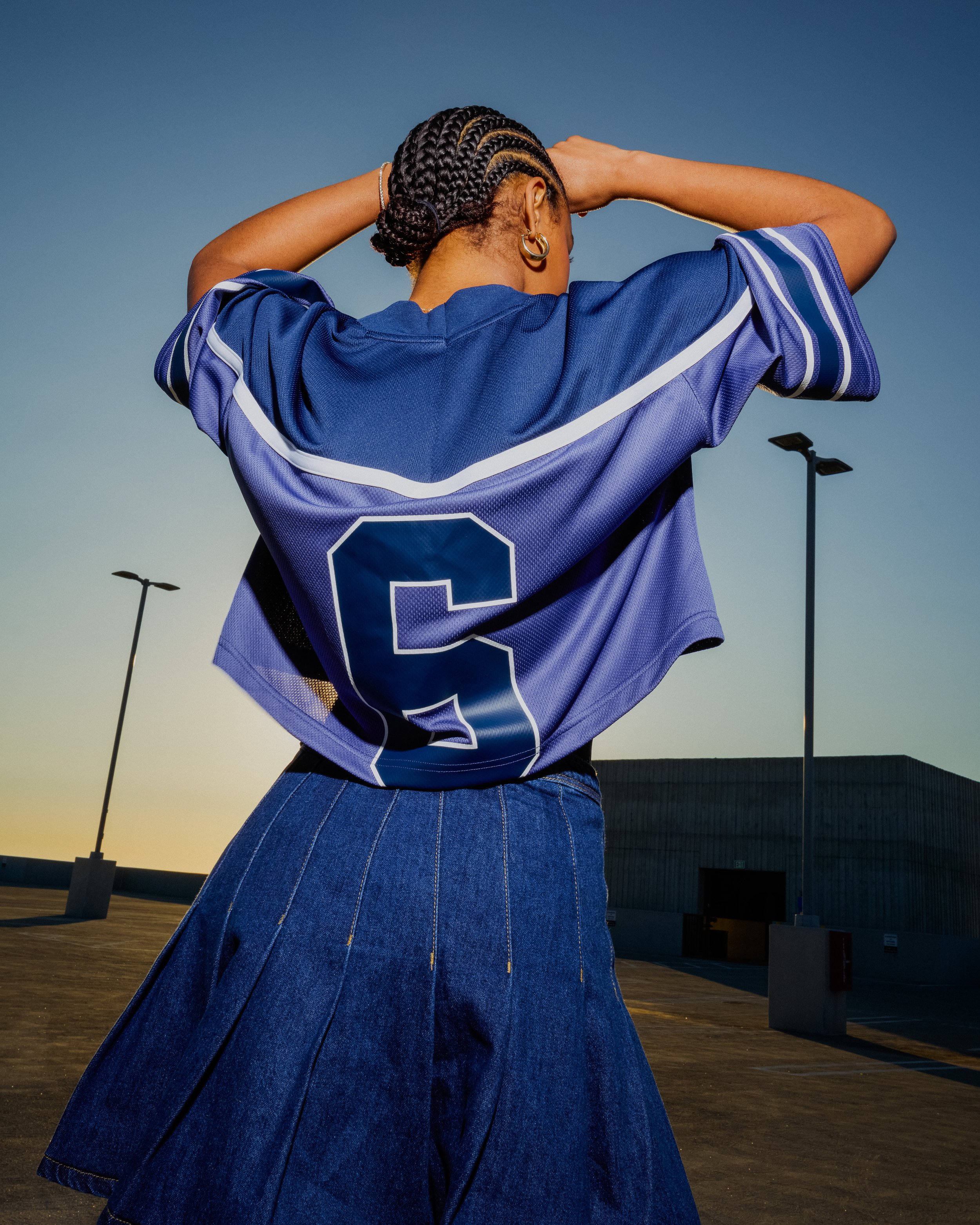 Back view of a woman with braided hair, wearing a blue sports jersey with the number 6, and a denim skirt, standing outdoors during sunset or dusk, with parking lot lamps and a building in the background.