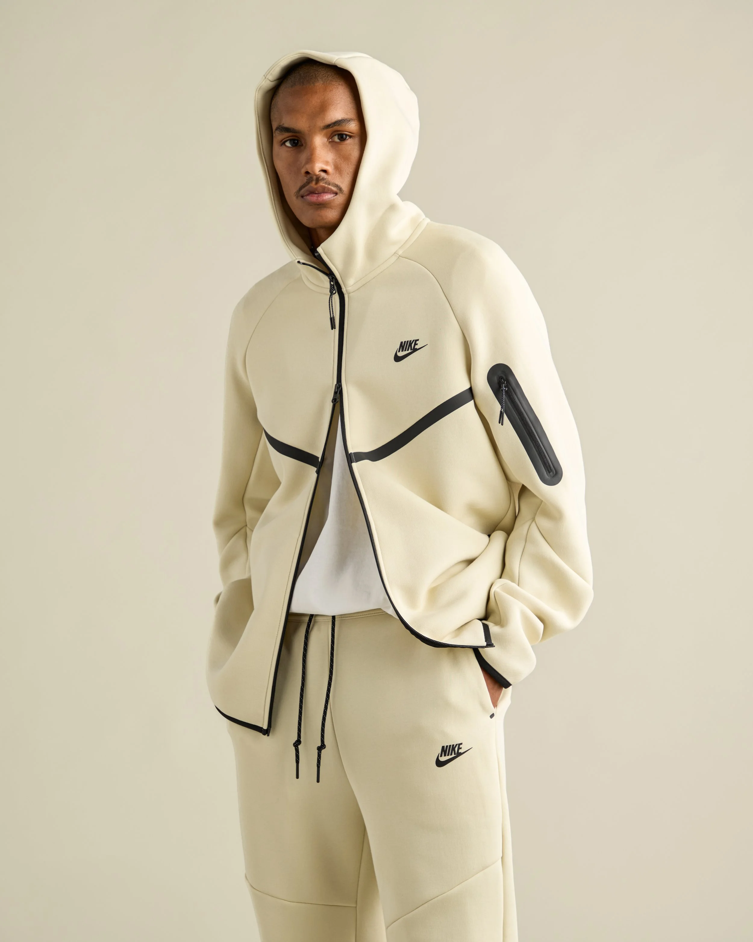 A young man with dark skin wearing a beige Nike hoodie with black accents and matching beige sweatpants with the Nike logo, standing against a plain beige background.