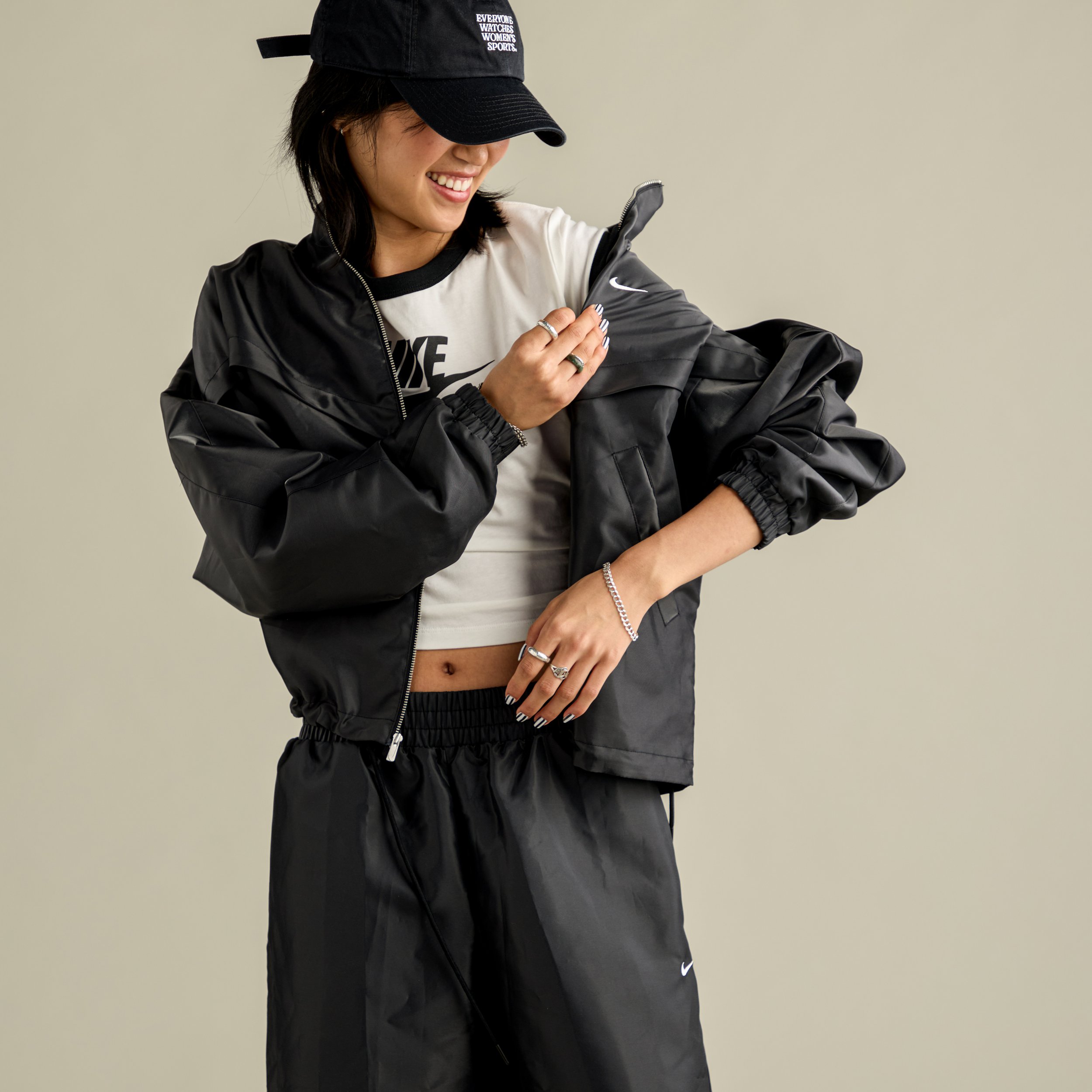 Woman wearing black Nike sportswear, including a cap, jacket, and pants, smiling and adjusting her jacket.