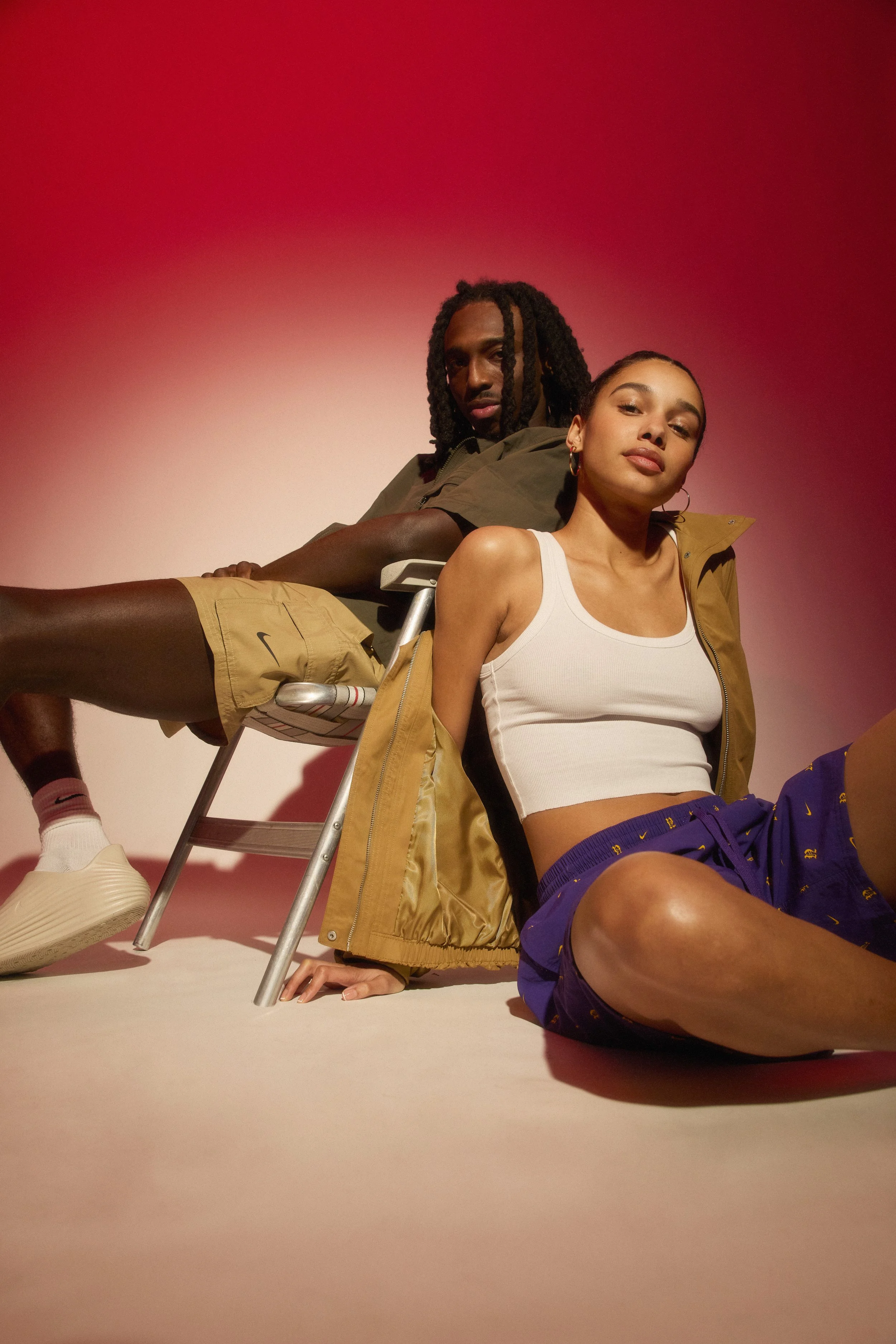 A young man and woman sitting on the floor back-to-back, against a pink gradient background. The man is wearing a dark green shirt and beige shorts, while the woman is wearing a white crop top and purple shorts.