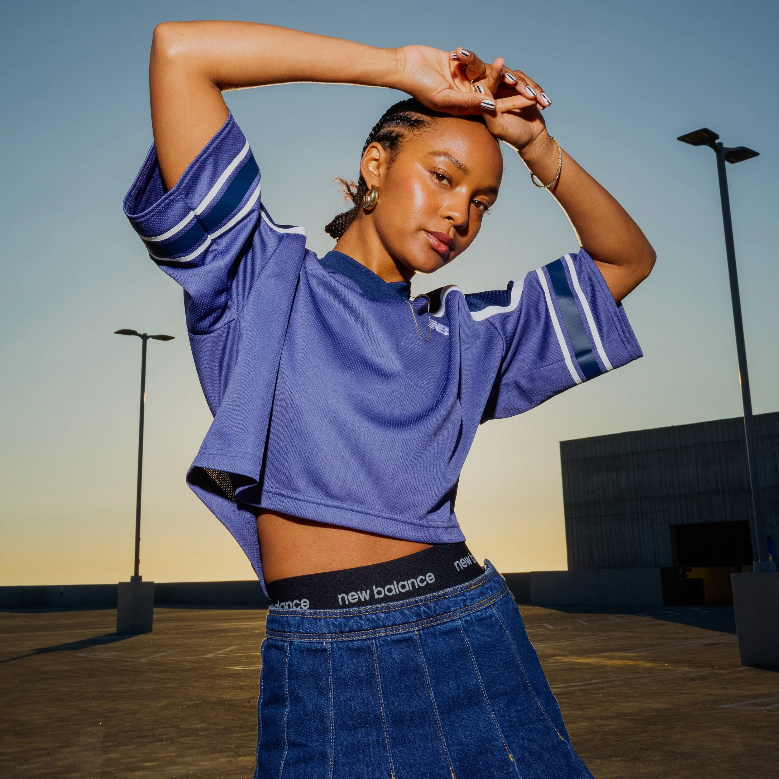 A woman in a blue sports jersey and denim skirt posing outdoors at sunset.