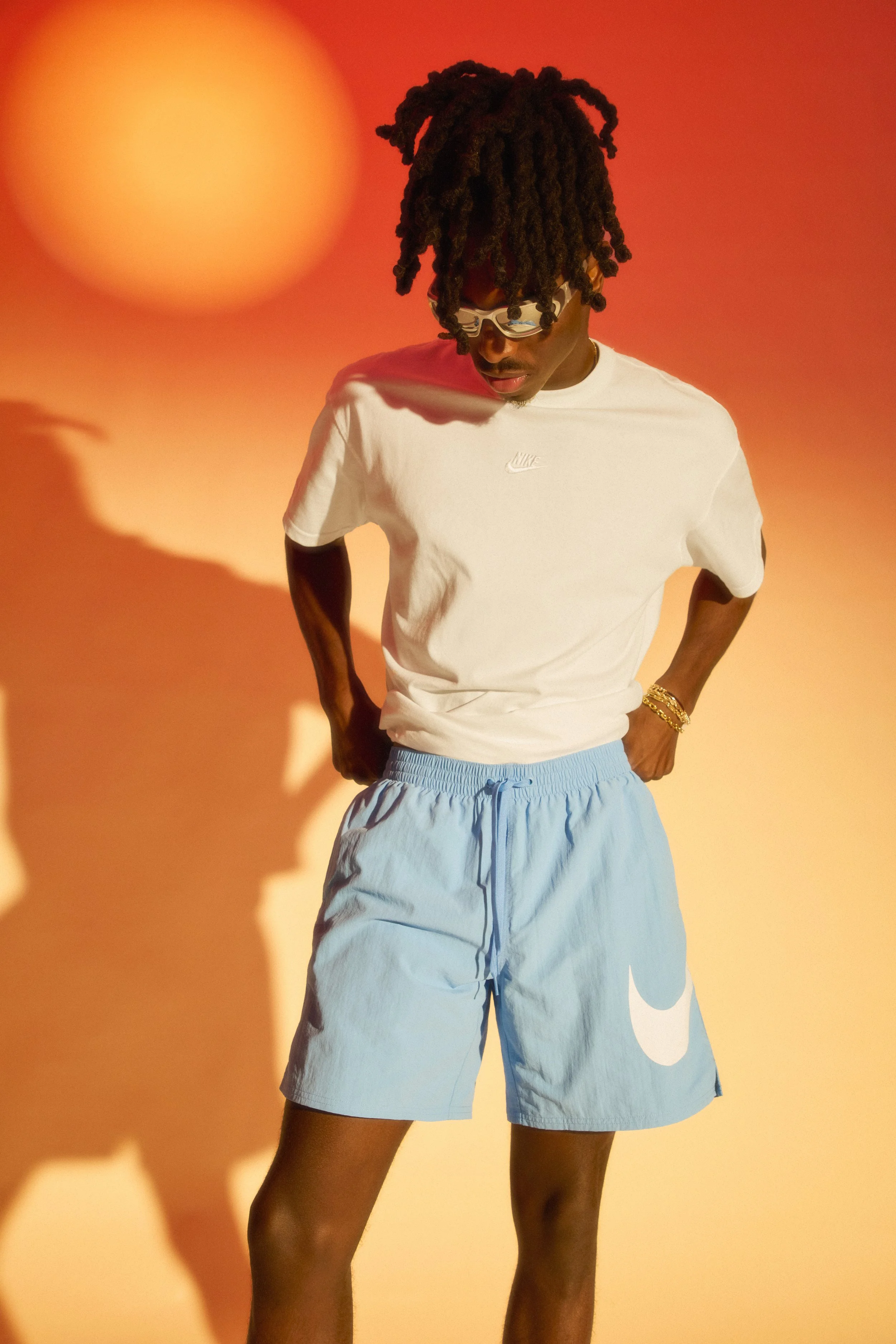 A young man with dreadlocks wearing sunglasses, a white Nike t-shirt, and light blue Nike shorts with a large white swoosh logo stands against an orange background with shadow patterns.