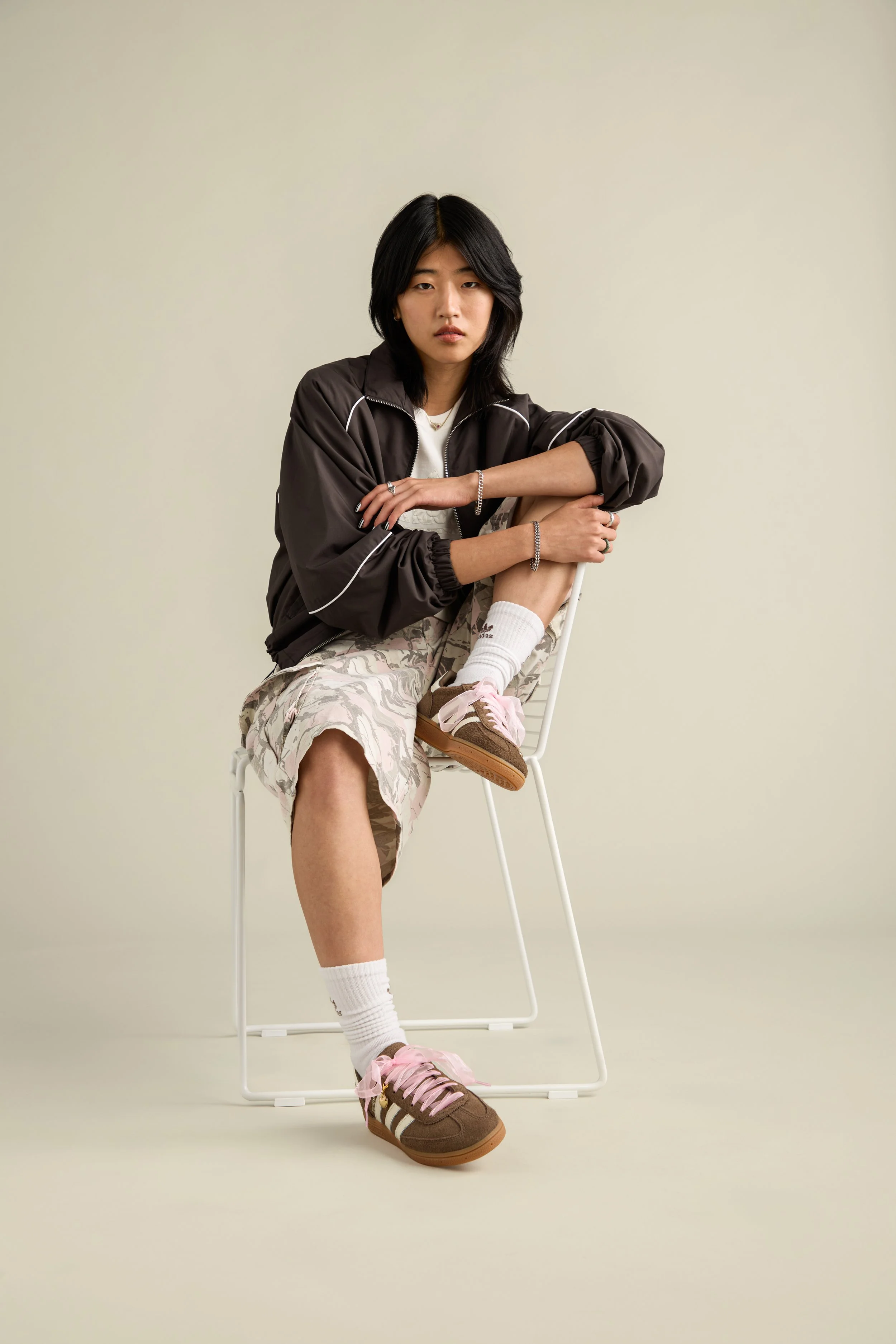 A young woman with black hair sitting on a white chair with her arms resting on her knee, wearing a black jacket, beige patterned shorts, white socks, and brown sneakers with pink shoelaces against a plain beige background.