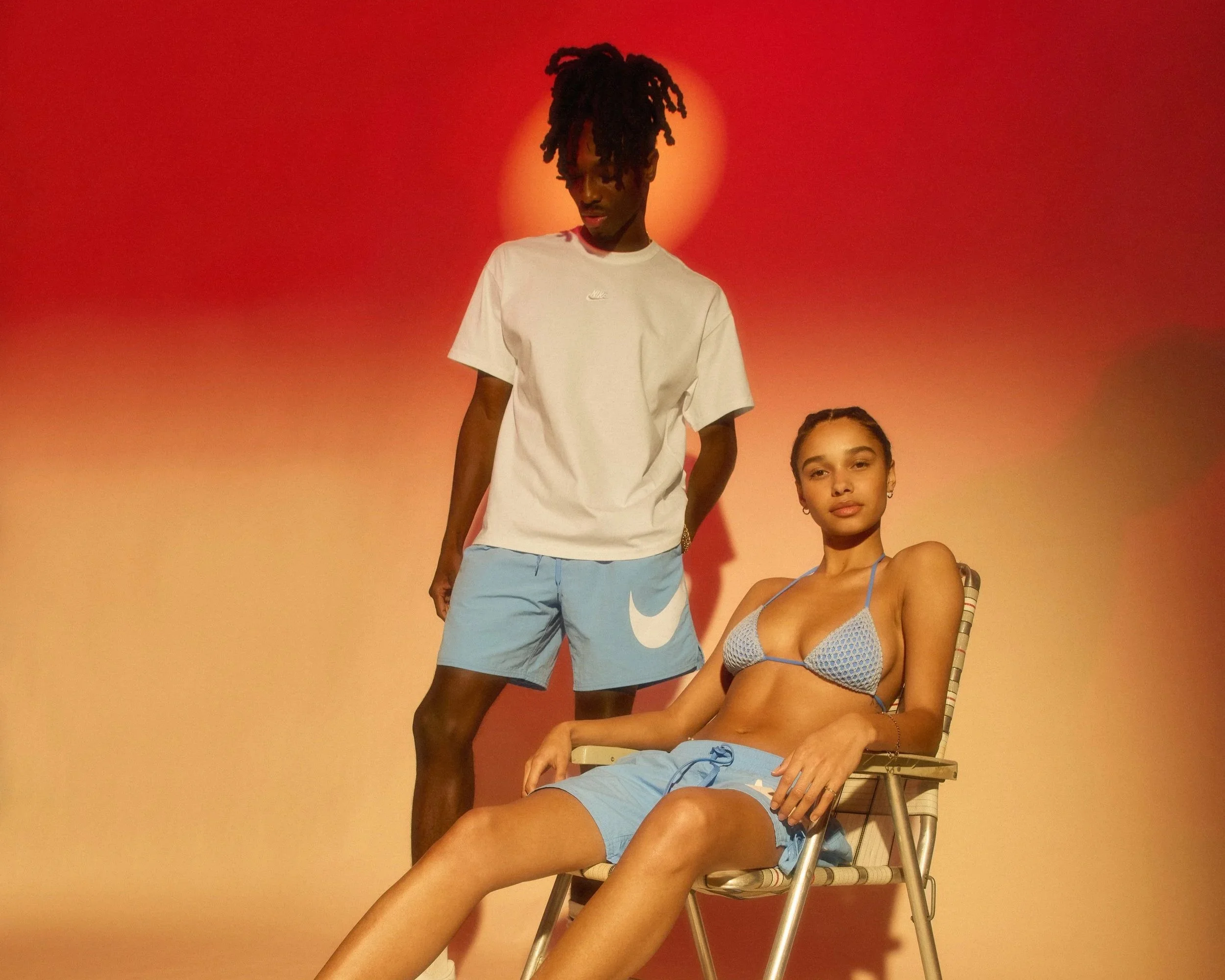 A young man standing and a young woman sitting on a chair against a colorful background with a glowing red-orange orb behind.