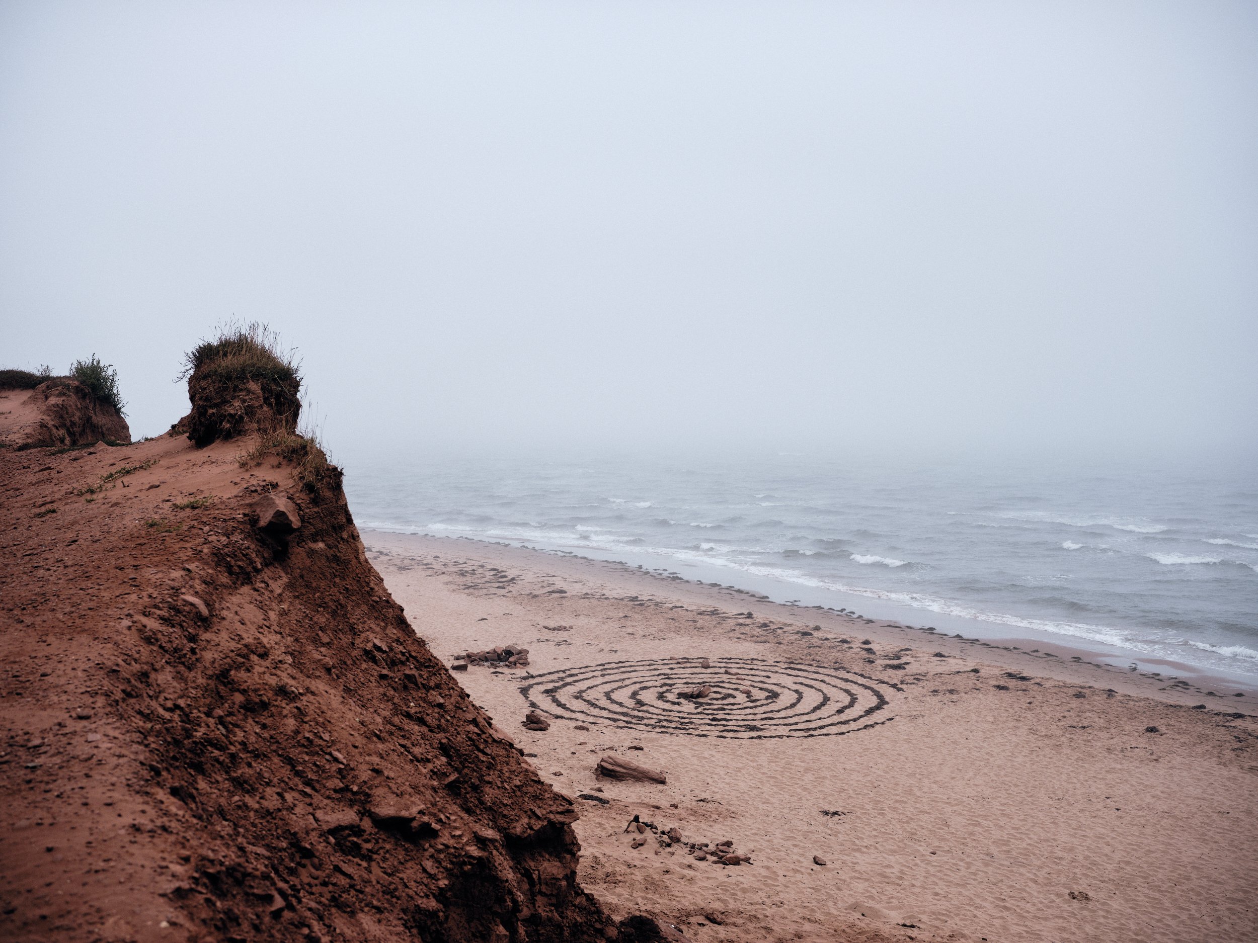 A foggy beach scene with eroded sandy cliffs on the left and the ocean in the background. A spiral pattern is drawn in the sand near the shoreline.