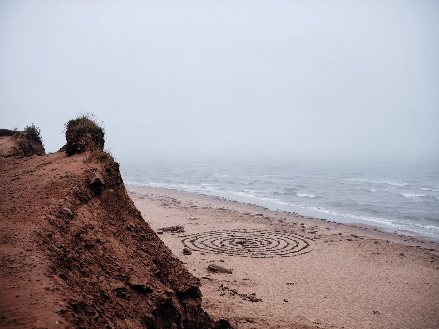 A foggy beach scene with eroded sandy cliffs on the left, an empty sandy shore, and waves in the ocean in the background. There is a spiral pattern drawn in the sand on the beach.