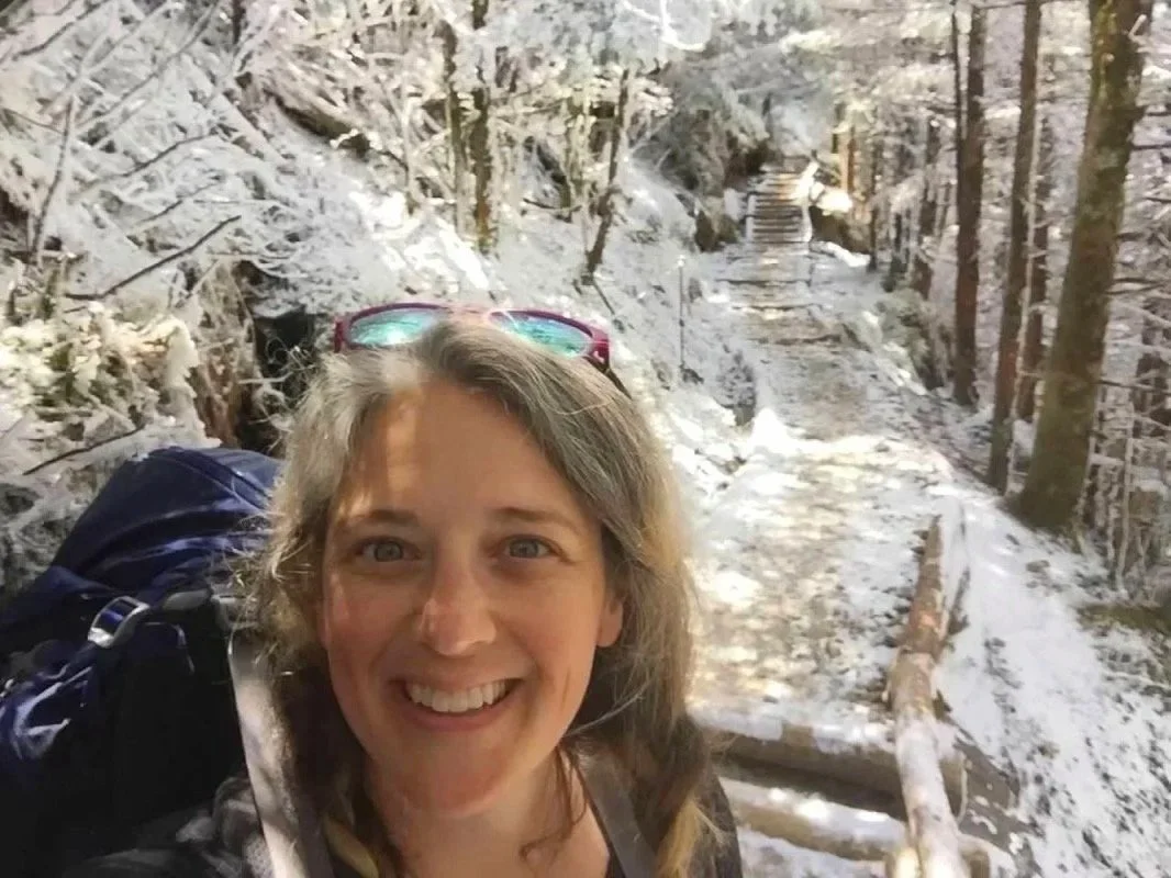 Woman taking a selfie on a snowy forest trail with trees and steps in the background.