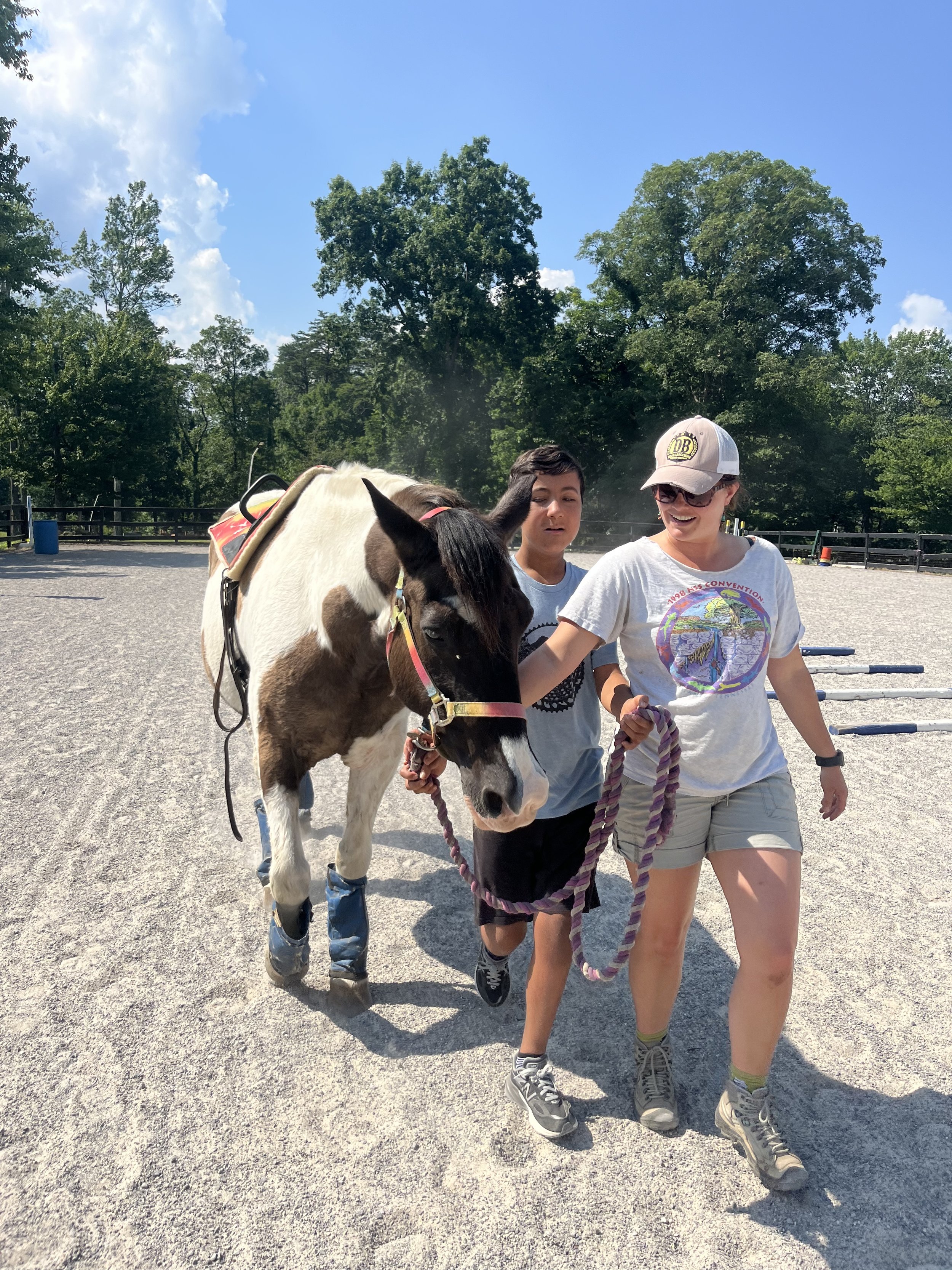 Two women and a young boy walking a pony on a leash in an outdoor riding arena with trees and a blue sky in the background.