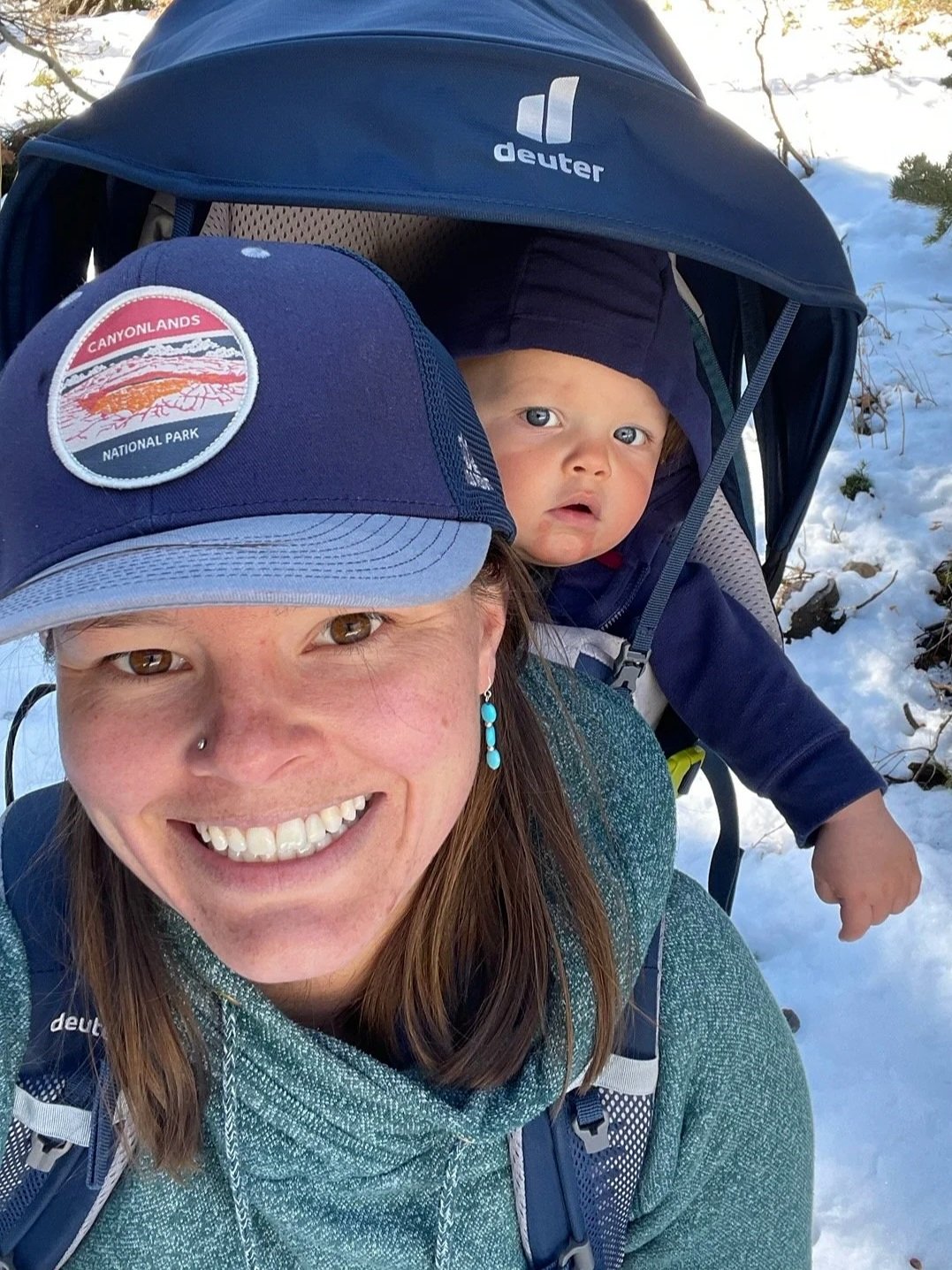 A woman smiling with a blue cap that has a patch reading 'CANYONLANDS NATIONAL PARK', on a snowy trail with a baby in a stroller, surrounded by snow and evergreen trees.