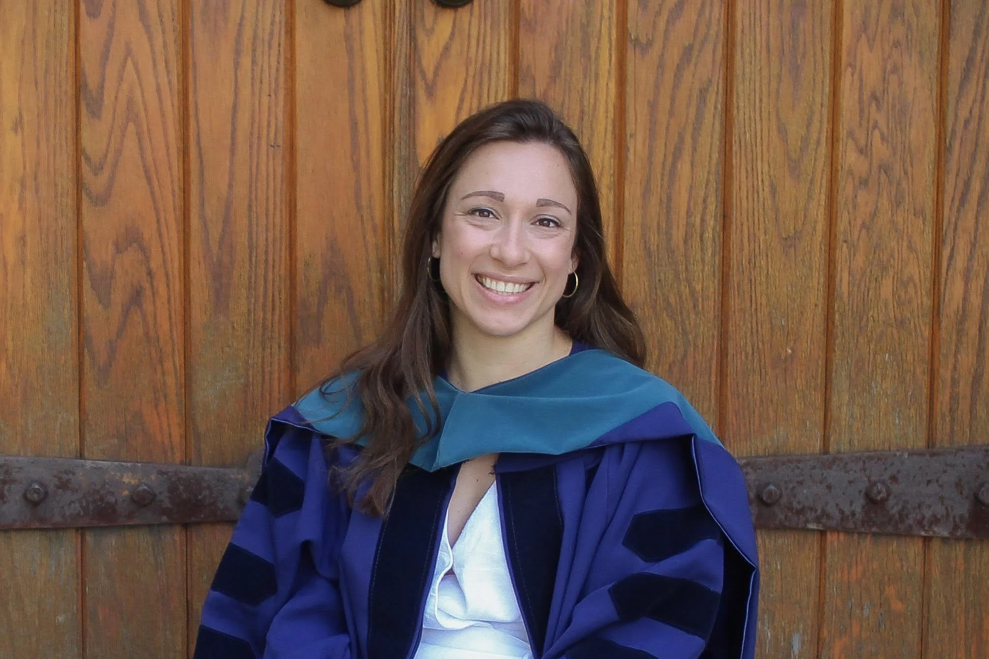 A woman with brown hair, glasses, and a striped shirt smiling outdoors with a child in a tie-dye shirt.
