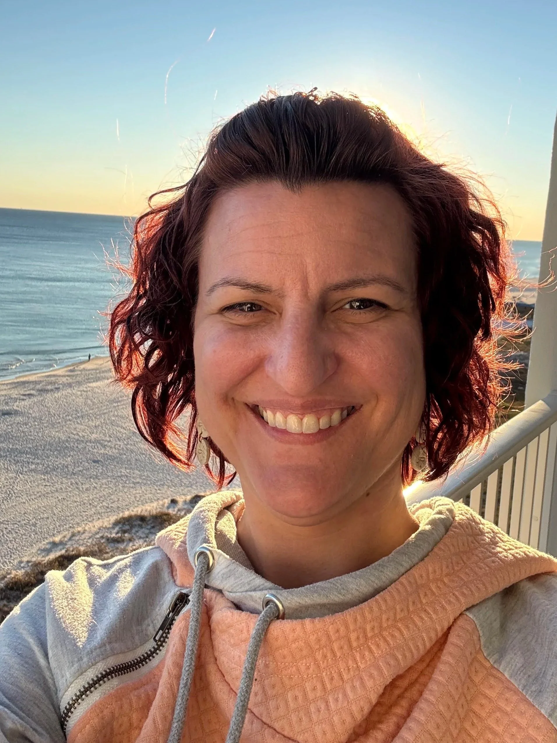 A woman smiling with short, curly red hair, standing on a beach at sunset, wearing a gray and peach hoodie, with the ocean and a yellow-orange sky in the background.