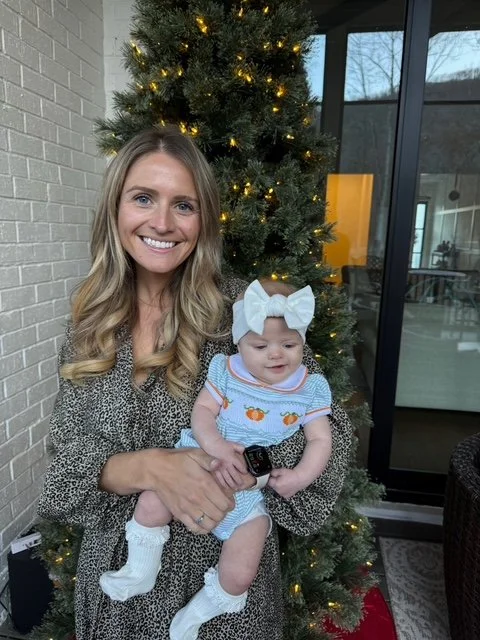 A woman with long blonde hair smiling while holding a baby girl with a large white bow on her head, in front of a decorated Christmas tree.