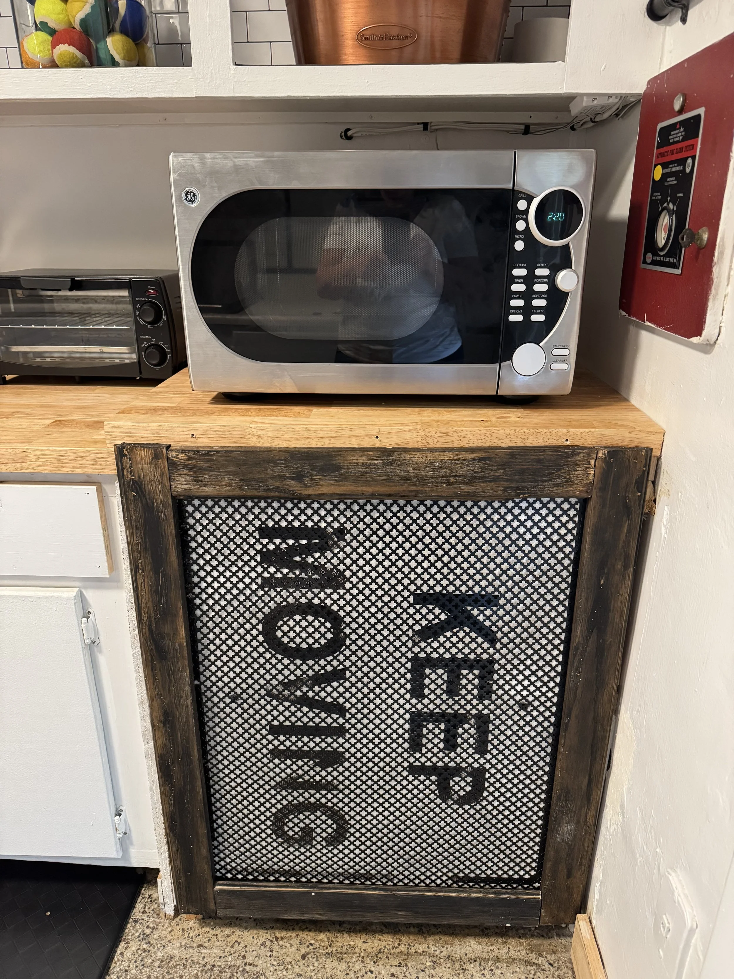 A stainless steel microwave oven on a wooden countertop with a black digital clock showing 2:20. There is a mesh panel below the microwave with the word 'LOVE' on it. To the left is a toaster oven, and to the right is a red fire alarm or control box 