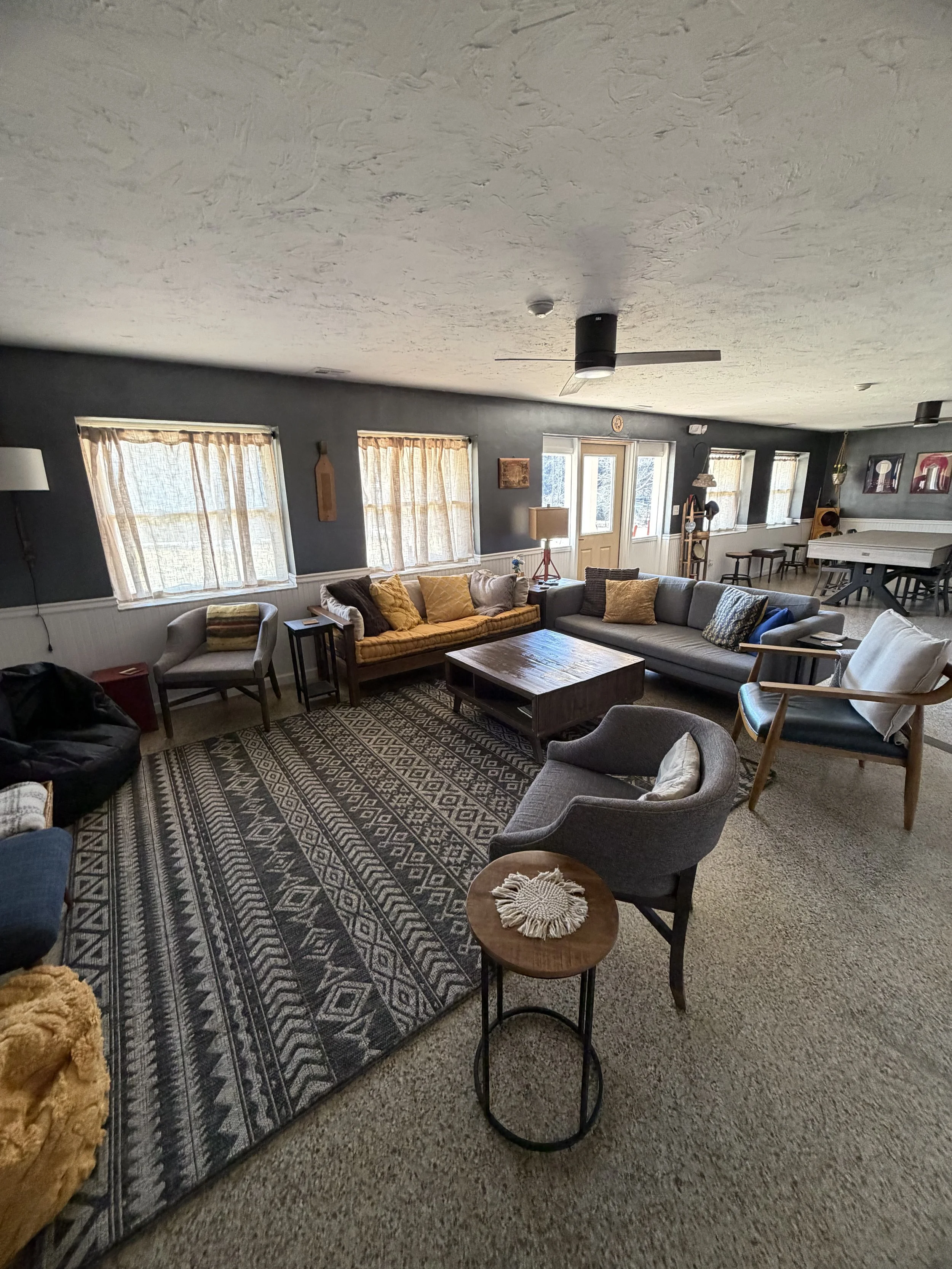 A spacious living room with multiple windows, couches, armchairs, a rug, and decorative items, with natural light coming in.