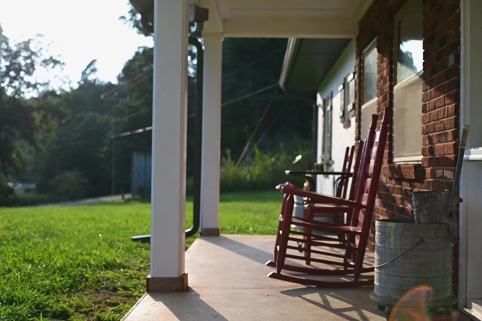 Front porch of a house with three red rocking chairs, a metal bucket, and a small side table, surrounded by green grass and trees in the background.