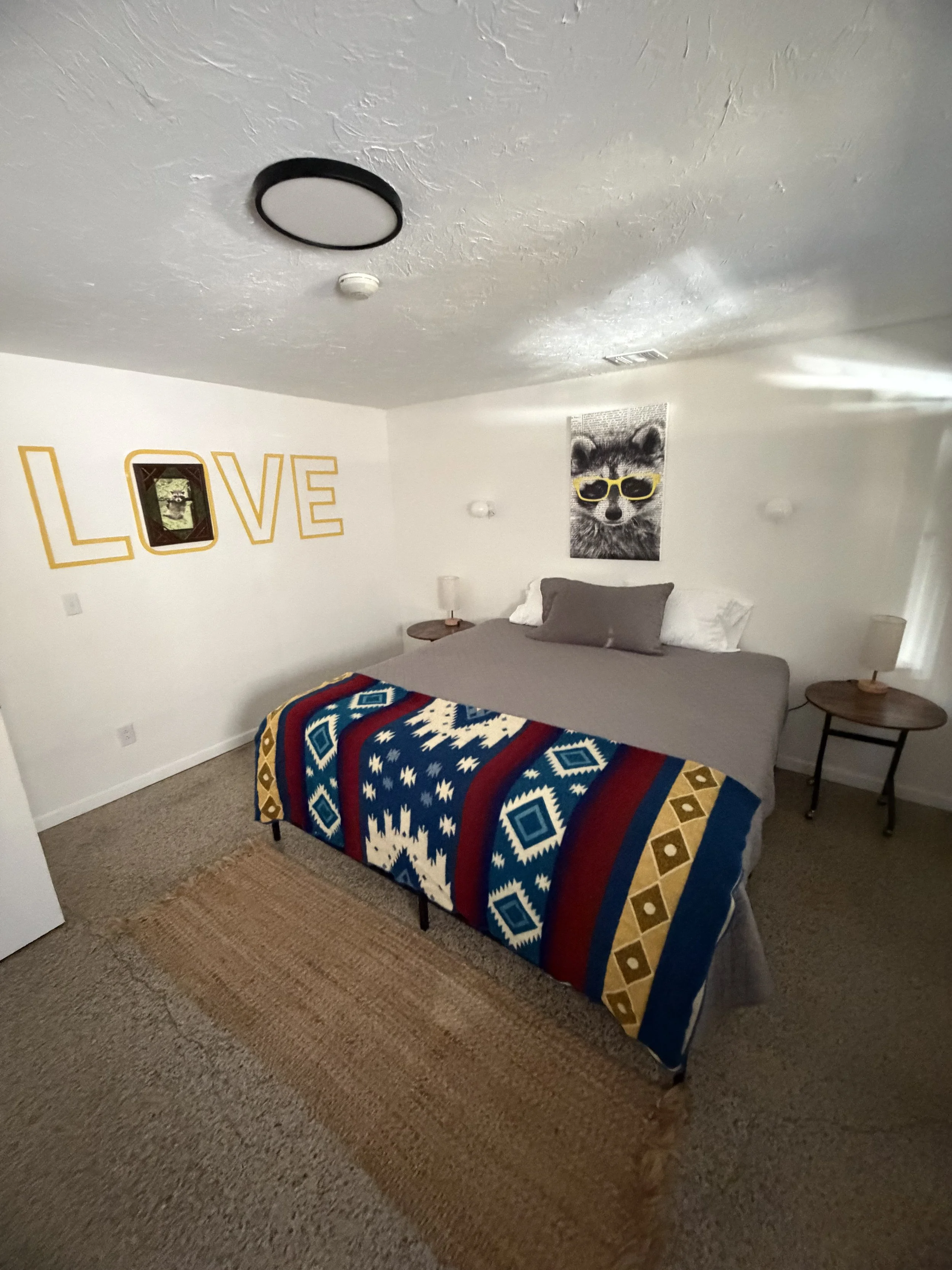 A bedroom with a bed covered in a blue, red, and white patterned blanket, gray and white pillows, and two wooden nightstands with lamps on each side. The wall behind the bed features a colorful poster of a raccoon wearing yellow sunglasses and a yell