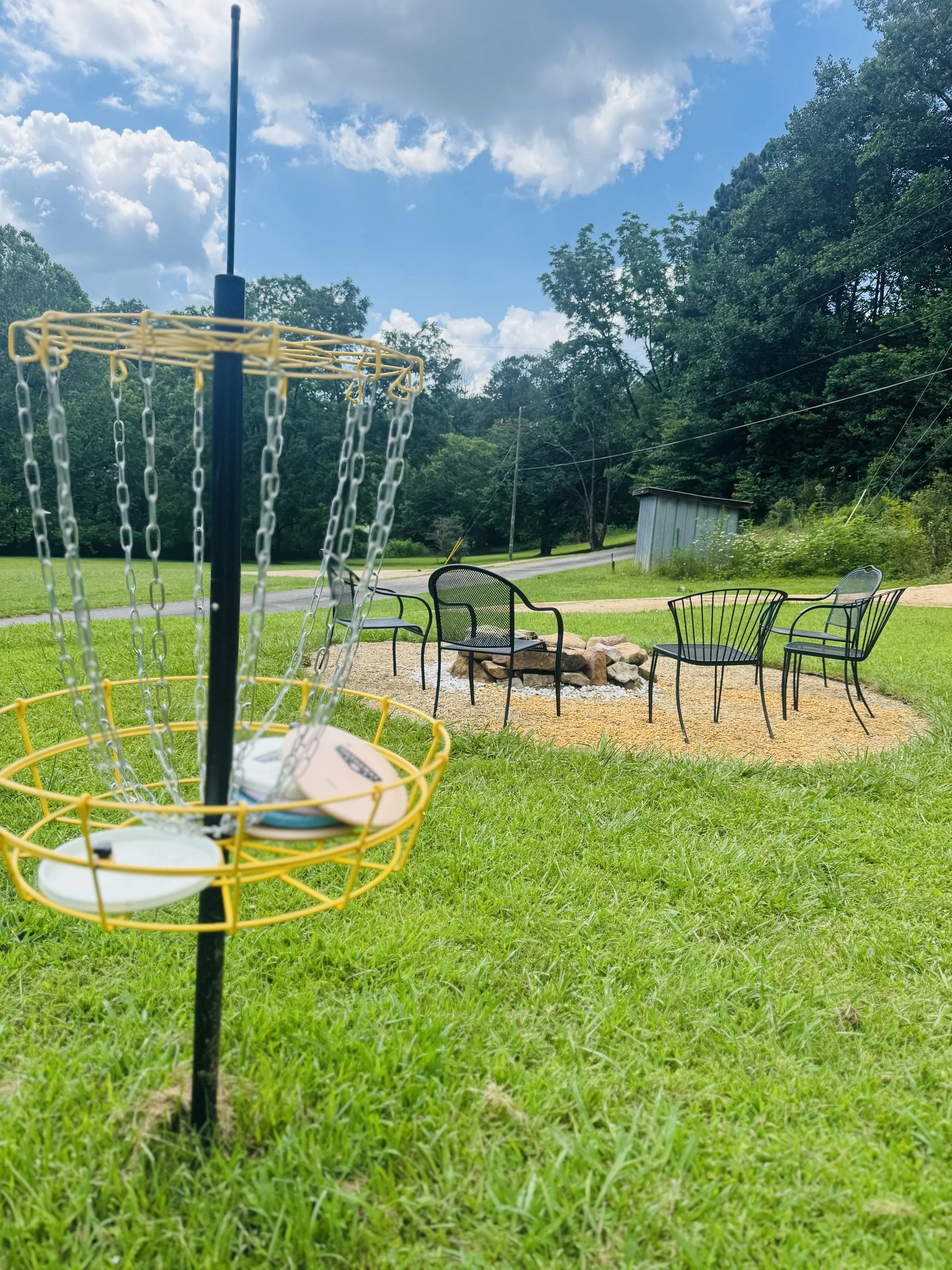 A disc golf basket with discs inside, positioned on green grass in a park. In the background, four black chairs are arranged around a fire pit with rocks, under a partly cloudy sky surrounded by trees.