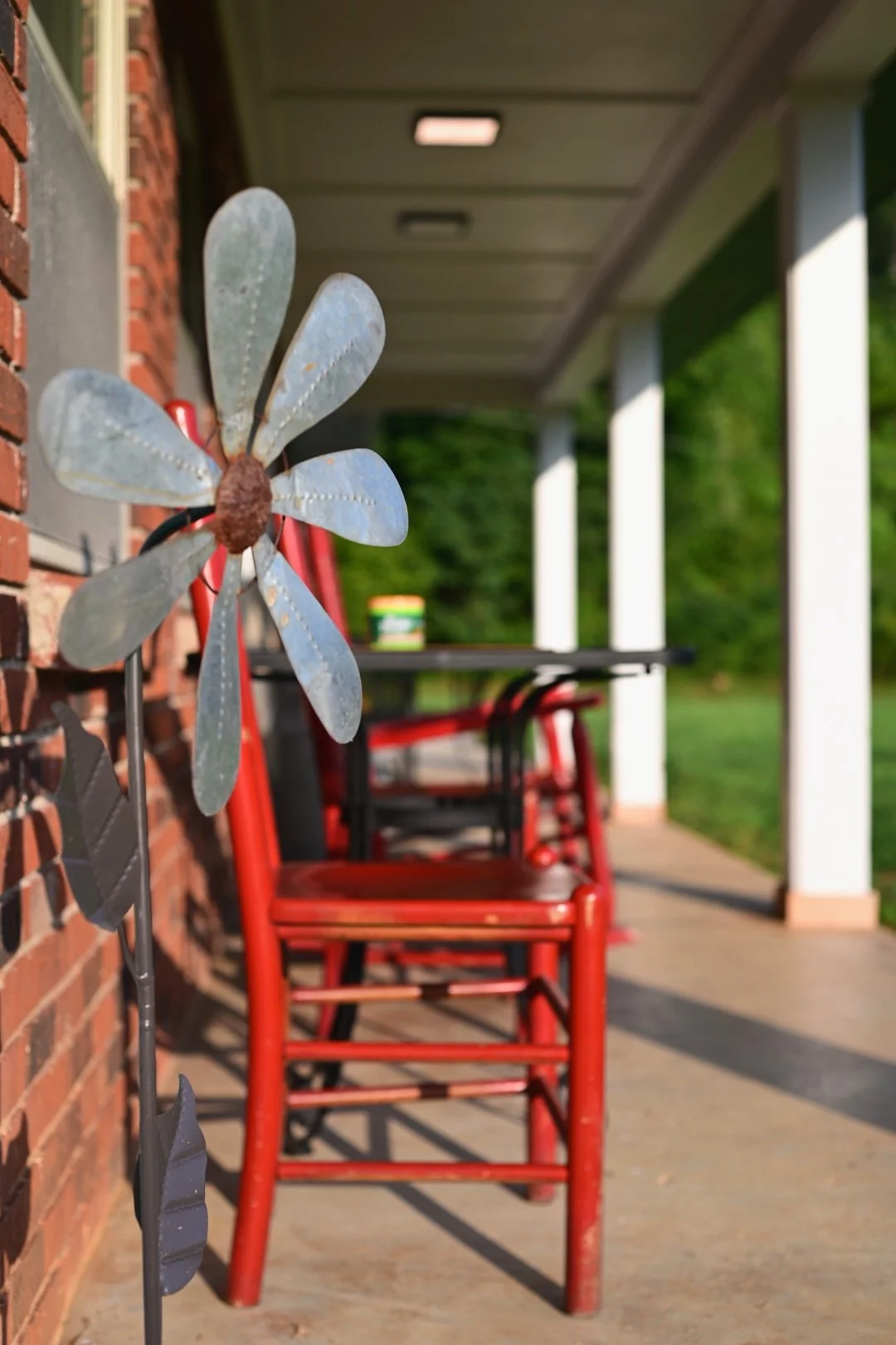Metal flower decoration on a porch with red chairs and a table, with greenery in the background.