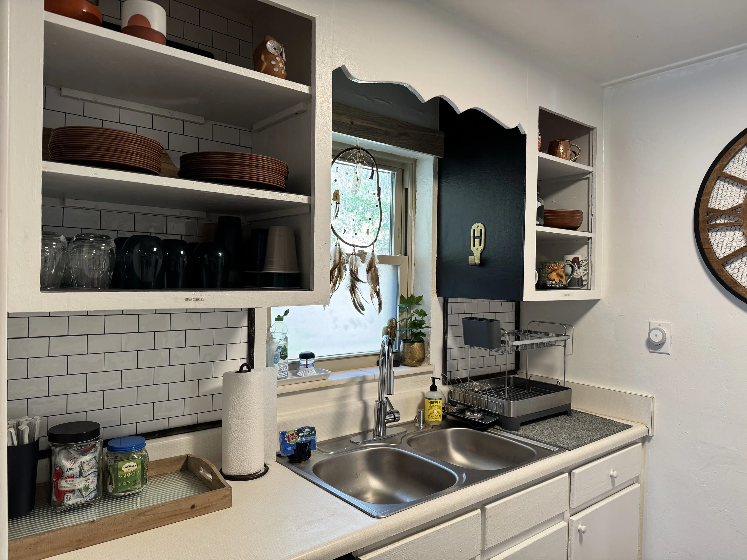 Kitchen with open shelves holding dishes, glasses, and cups above the sink. There is a small window with decorative hanging and a potted plant. The countertop has a paper towel roll, a jar of tea, and a tray with snack packs. A dish drying rack and v