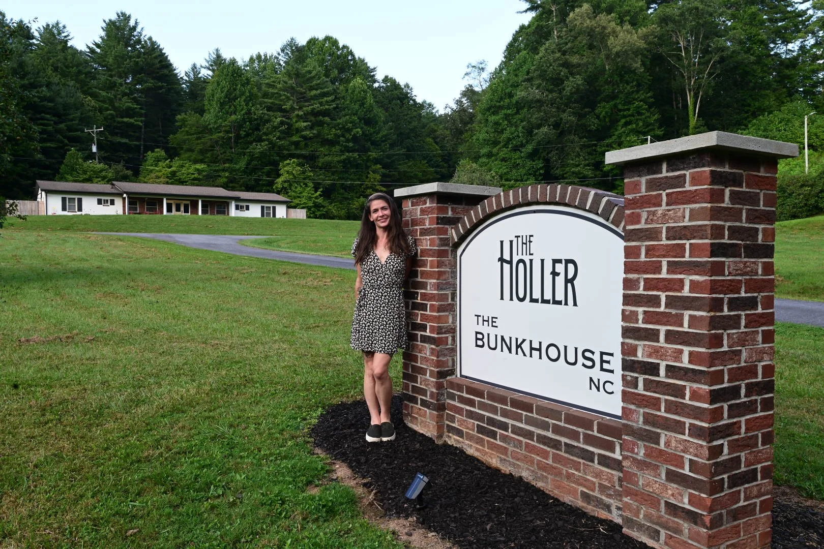 A woman in a black and white dress standing next to a brick sign that reads 'The Holler, The Bunkhouse NC' in a grassy area with a road and trees in the background.