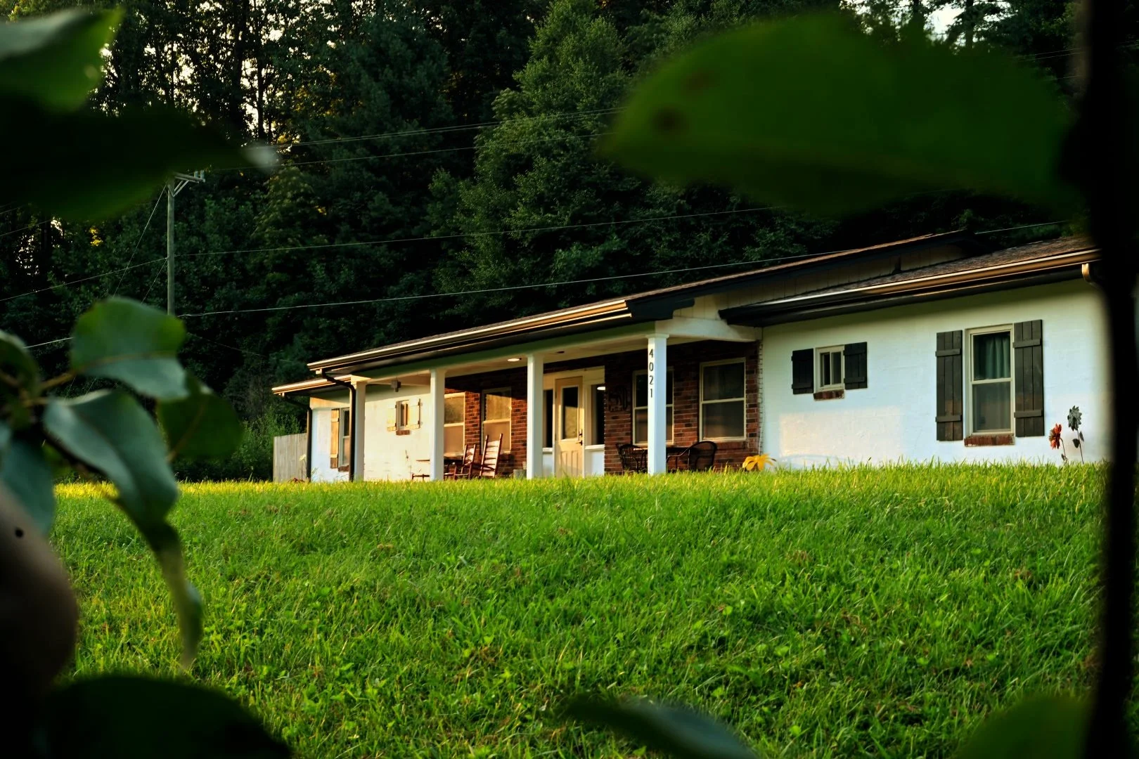 Front view of a single-story house with a porch and black shutters, surrounded by green grass and trees in the background.
