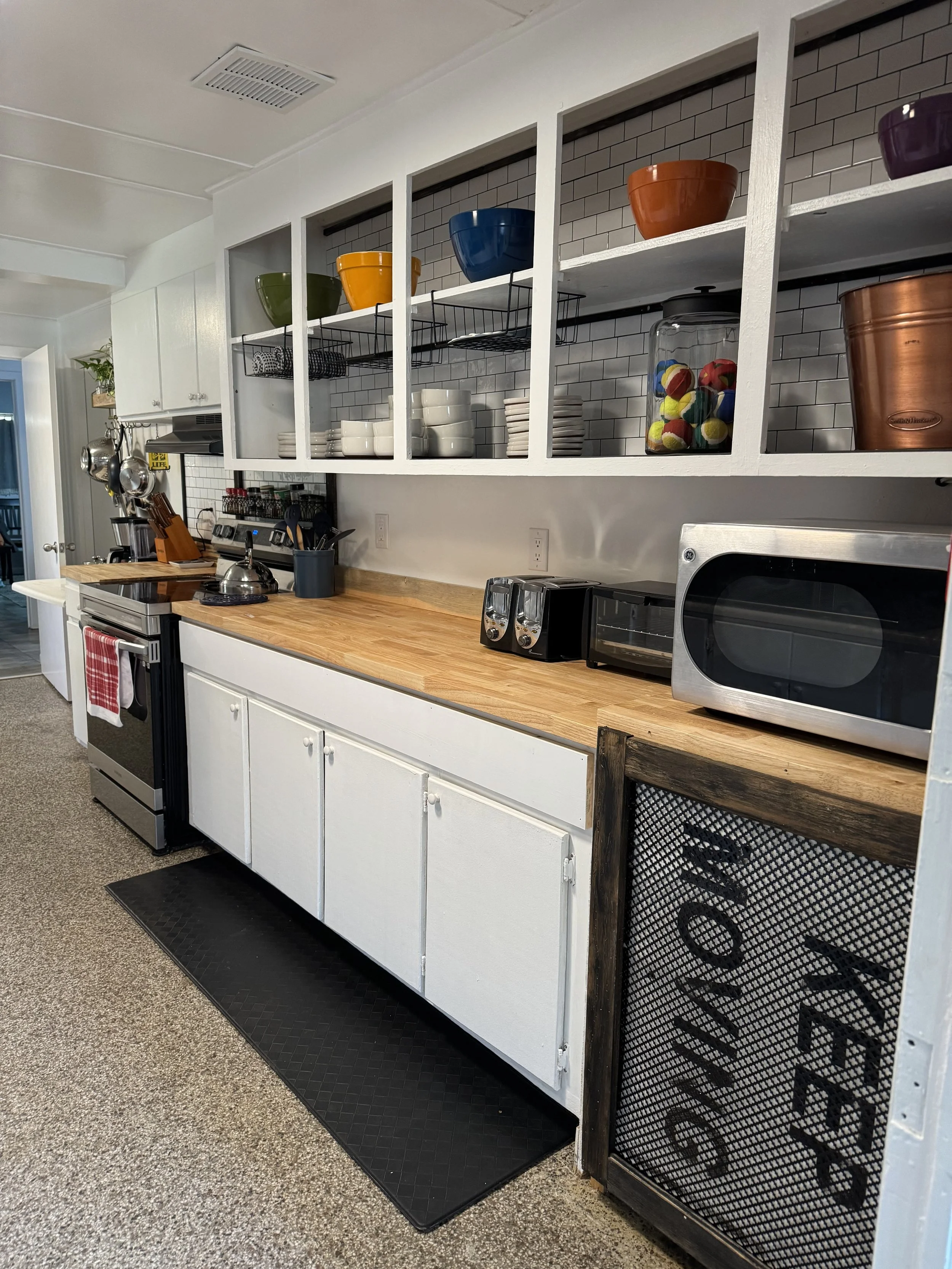 A kitchen with white cabinets, open shelving displaying colorful bowls, dishes, and a jar of tennis balls. There is a wood countertop with small appliances including toasters and a microwave. The kitchen has a stove, a black rug, and a black metal si