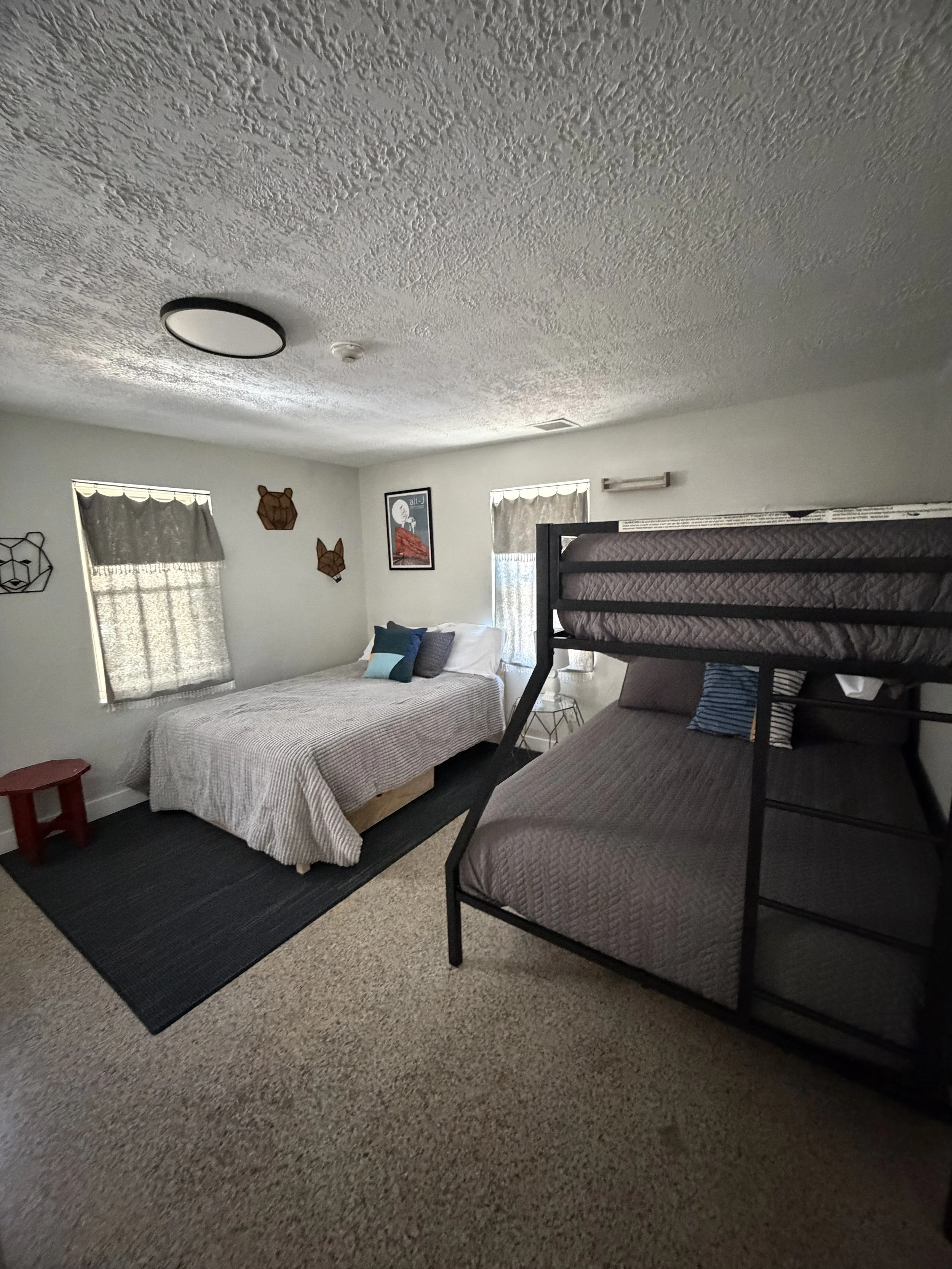 A bedroom with a bed, bunk bed, two windows with curtains, wall decorations, a small red stool, and a side table.