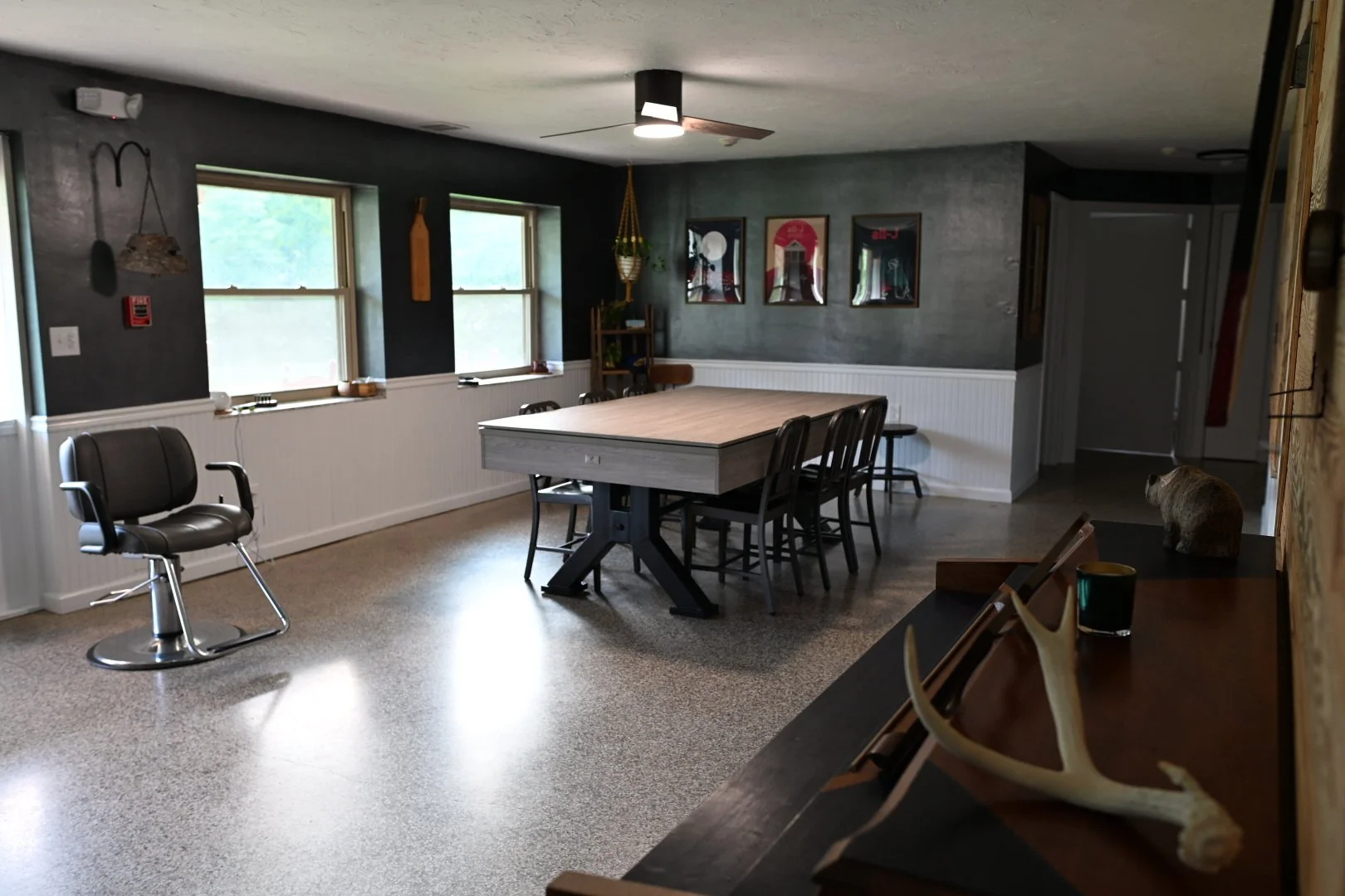 A spacious dining room with a large rectangular table, several black chairs, two windows with blinds, and decorative art on the walls. There is a bar stool to the left and a side counter with antlers, a mug, and a small animal figurine.