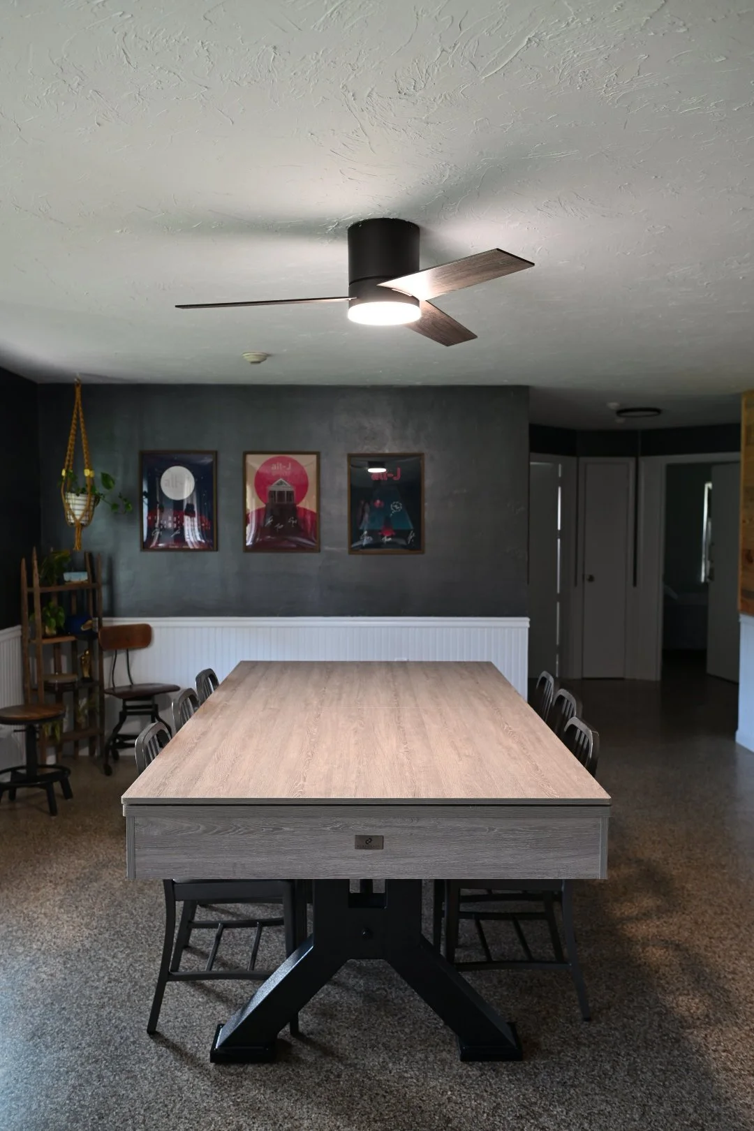 A dining room with a long wooden table and black chairs. There is a dark gray wall with three framed pictures, and a ceiling fan with wooden blades and a light. The room also has a small shelf with plants and some chairs in the corner.