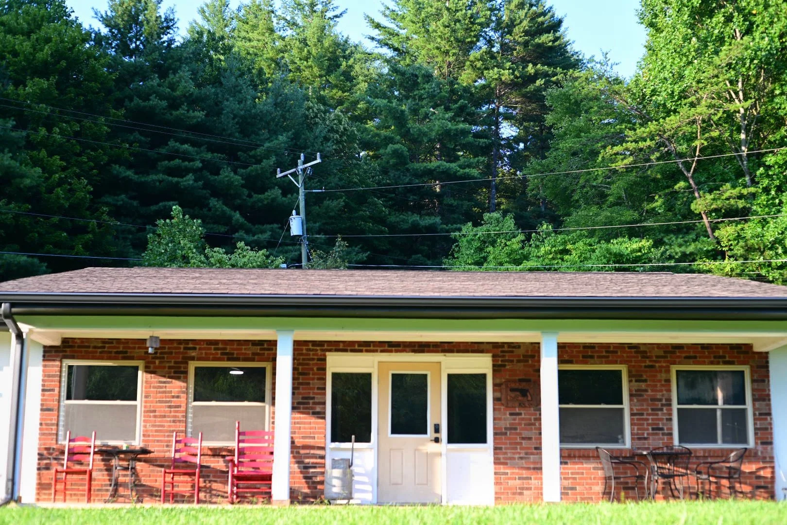 Single-story brick house with a covered porch, three windows with screens, and outdoor furniture including chairs and a table, set against a backdrop of tall, lush green trees.