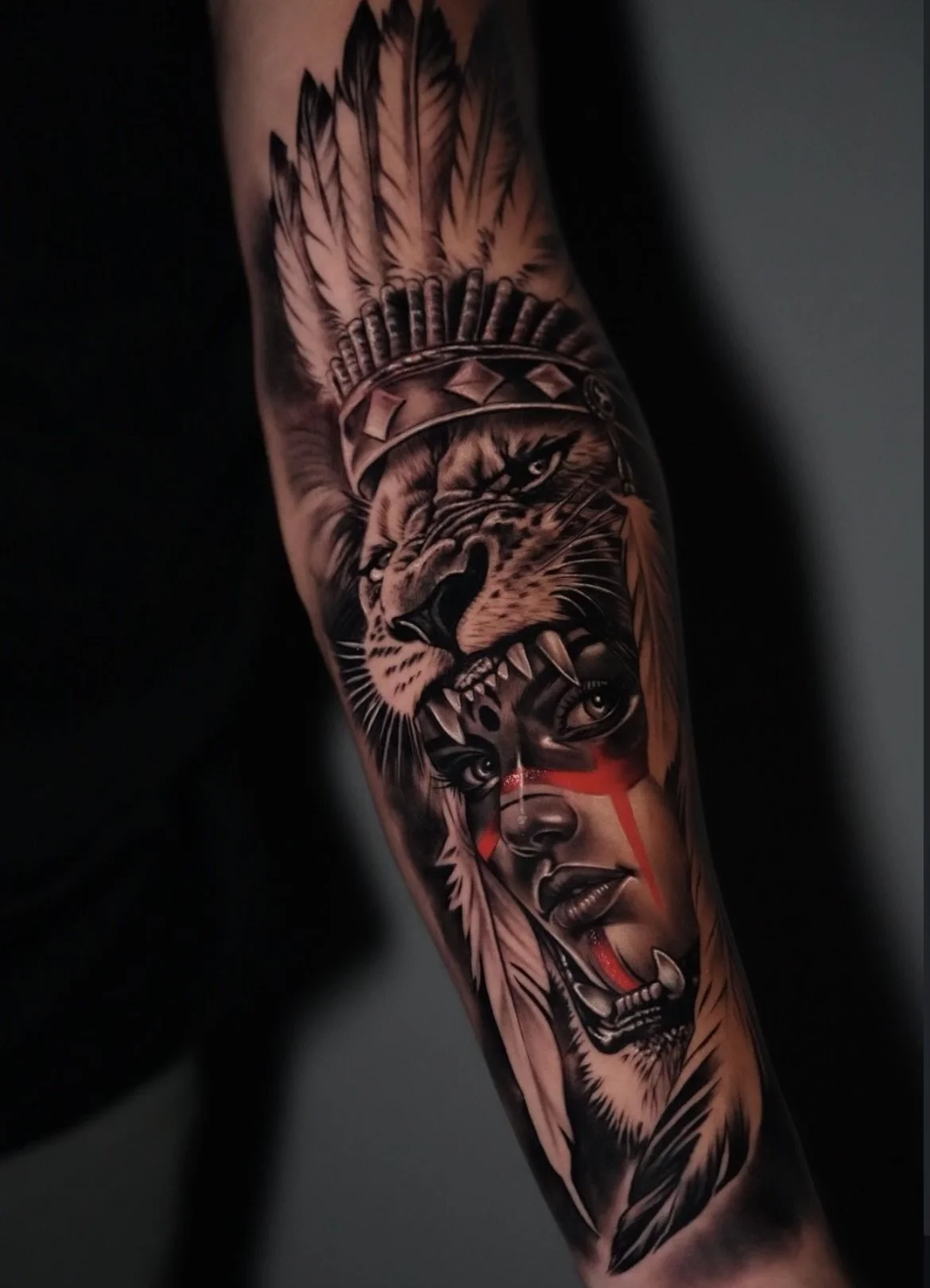 Tattoo of a fierce lion wearing a Native American headdress with feathers, combined with a woman's face with red war paint, on a person's arm.