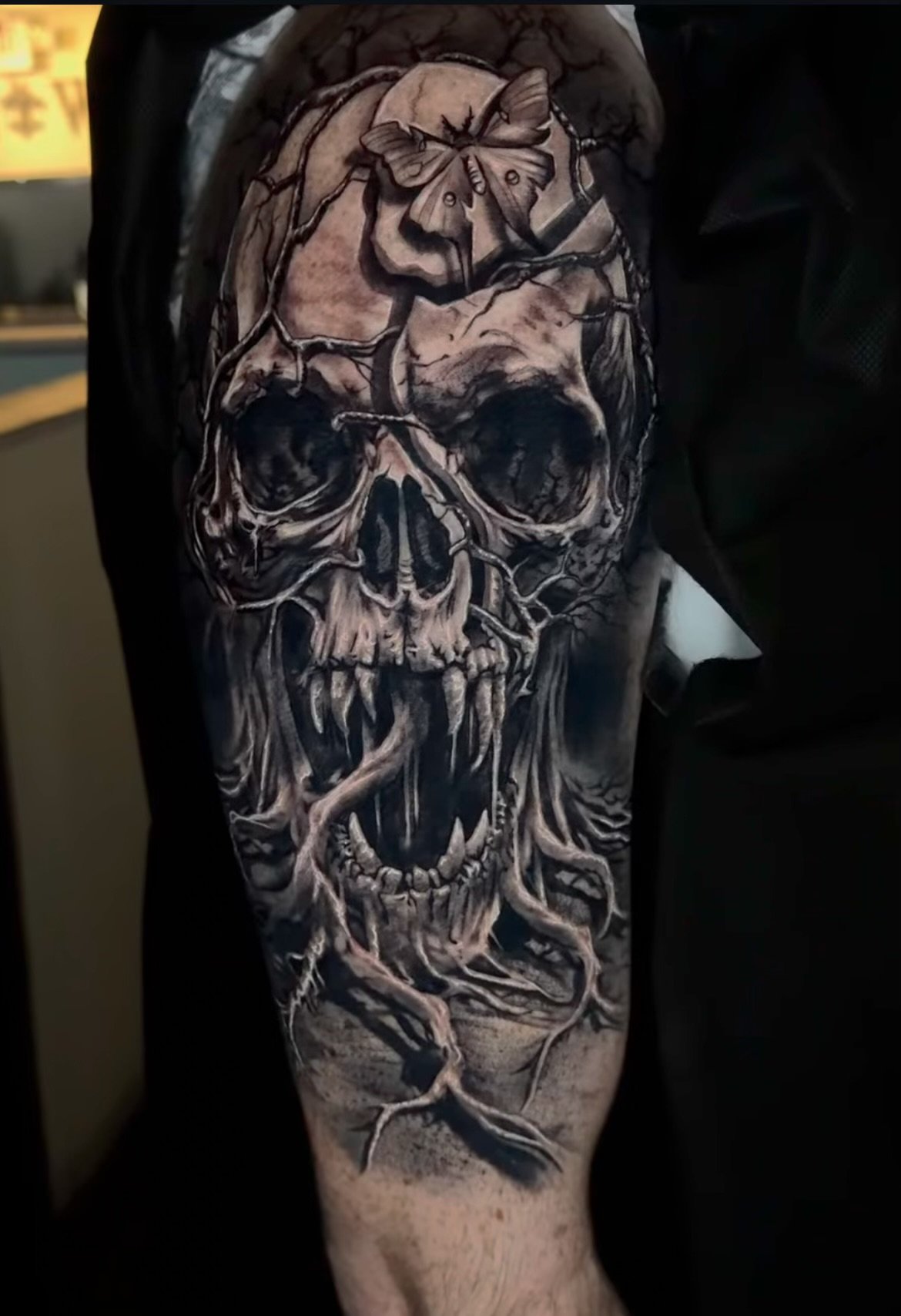 Black and gray tattoo of a skull with tree roots, a blooming flower, and a moth on the forehead on a person's arm.