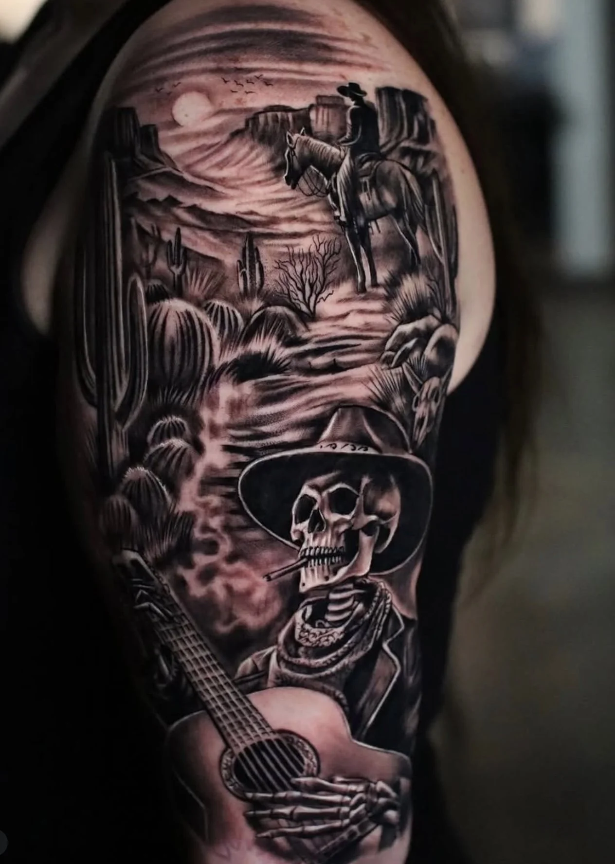 A black and gray tattoo of a cowboy with a hat, smoking a cigarette, standing beside a skeleton playing a guitar, with a desert landscape and full moon in the background.