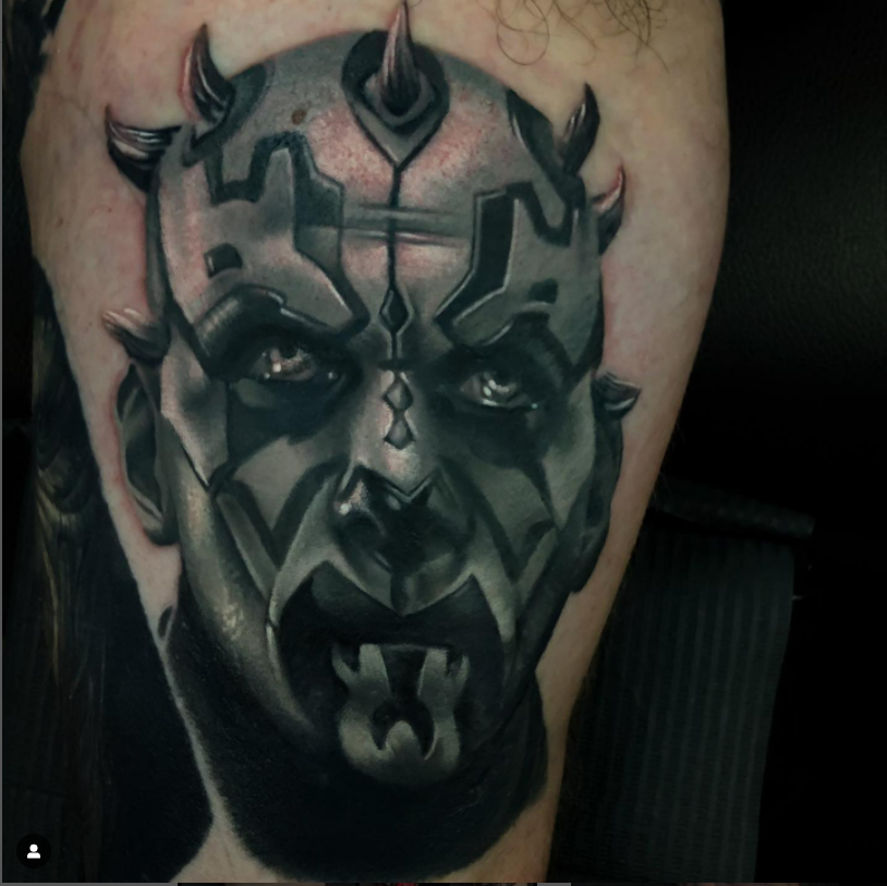 A black and gray tattoo of a fierce, armored face with animalistic features, including sharp horns and intense eyes.